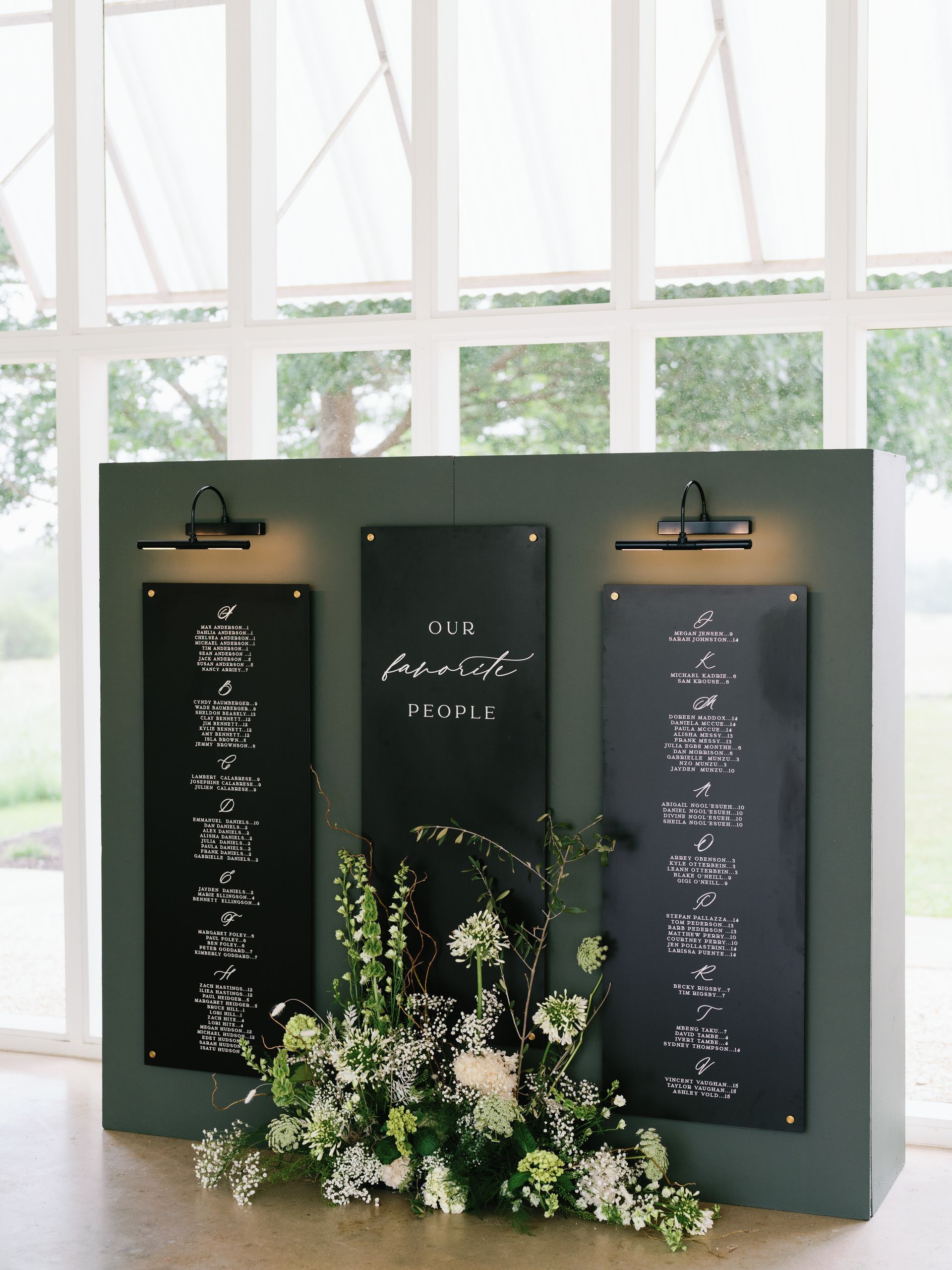 A wedding reception seating chart with three black panels and white text on them. There are long lamp hanging above the names as well as white and green flowers on the ground in front. 