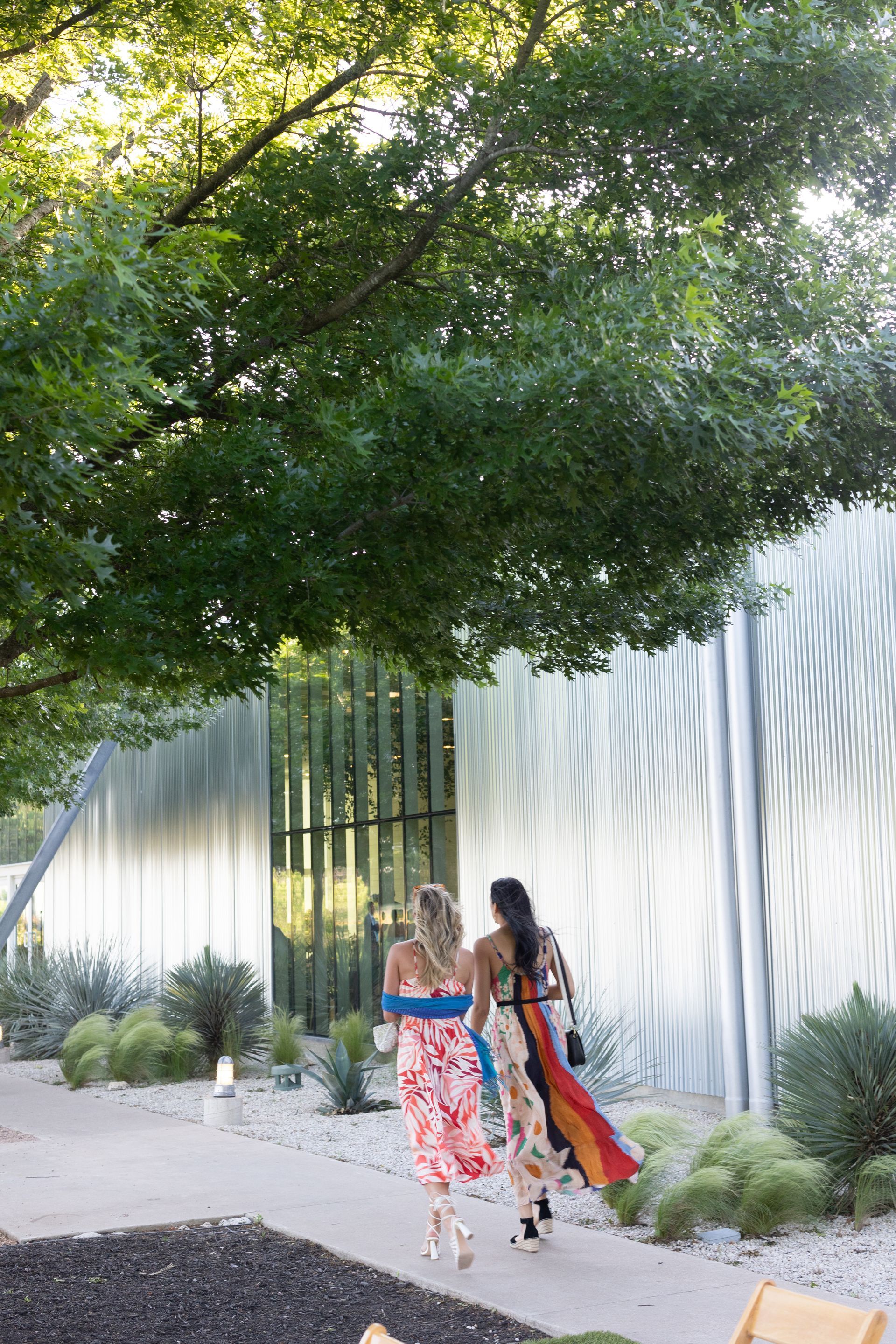 Two women walking on a path towards a modern, textured wall, under a tree.