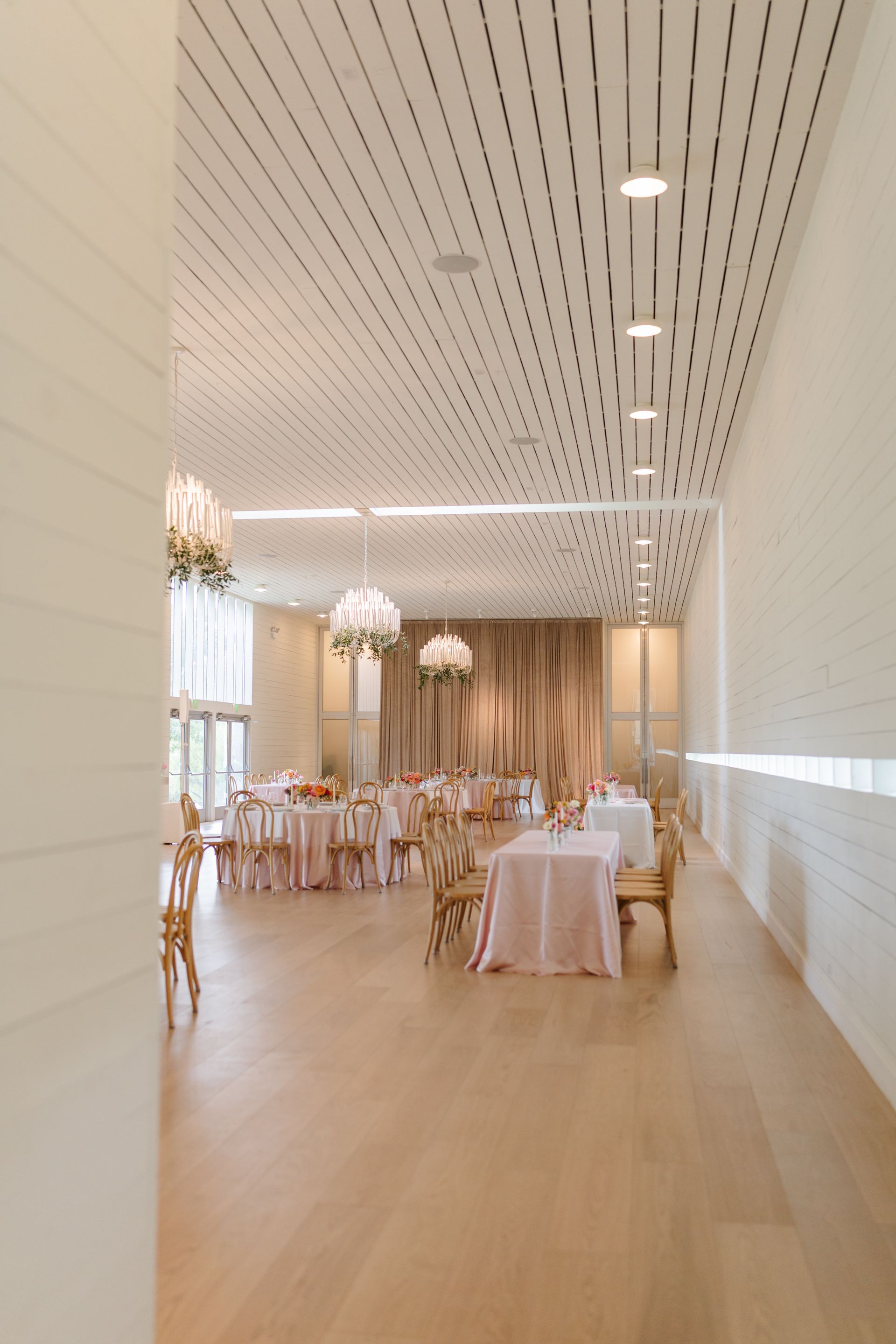 A light-filled event space with tables set for a wedding. Pale pink linens, wooden floors, and patterned ceiling.