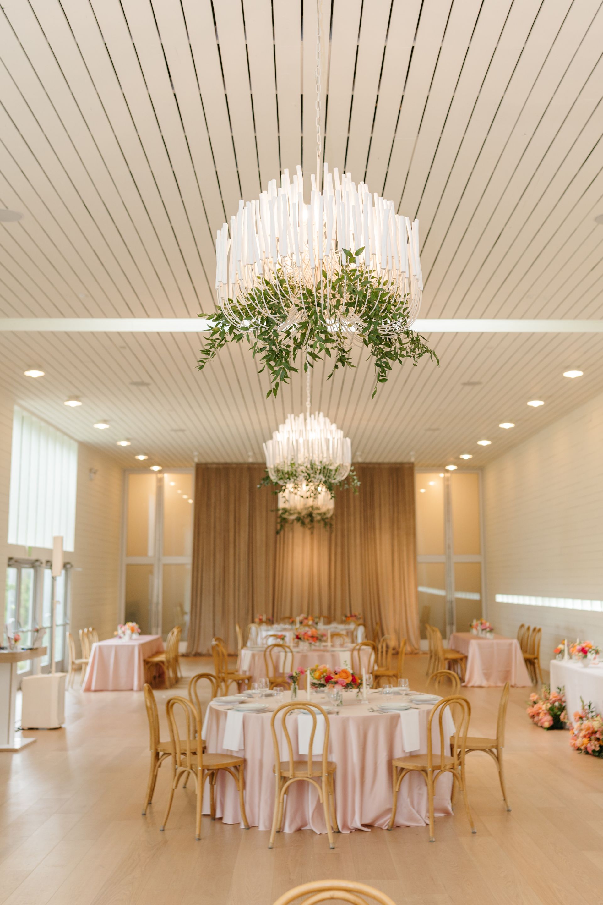 Elegant event space with round tables set for a reception, accented by chandeliers and floral arrangements.
