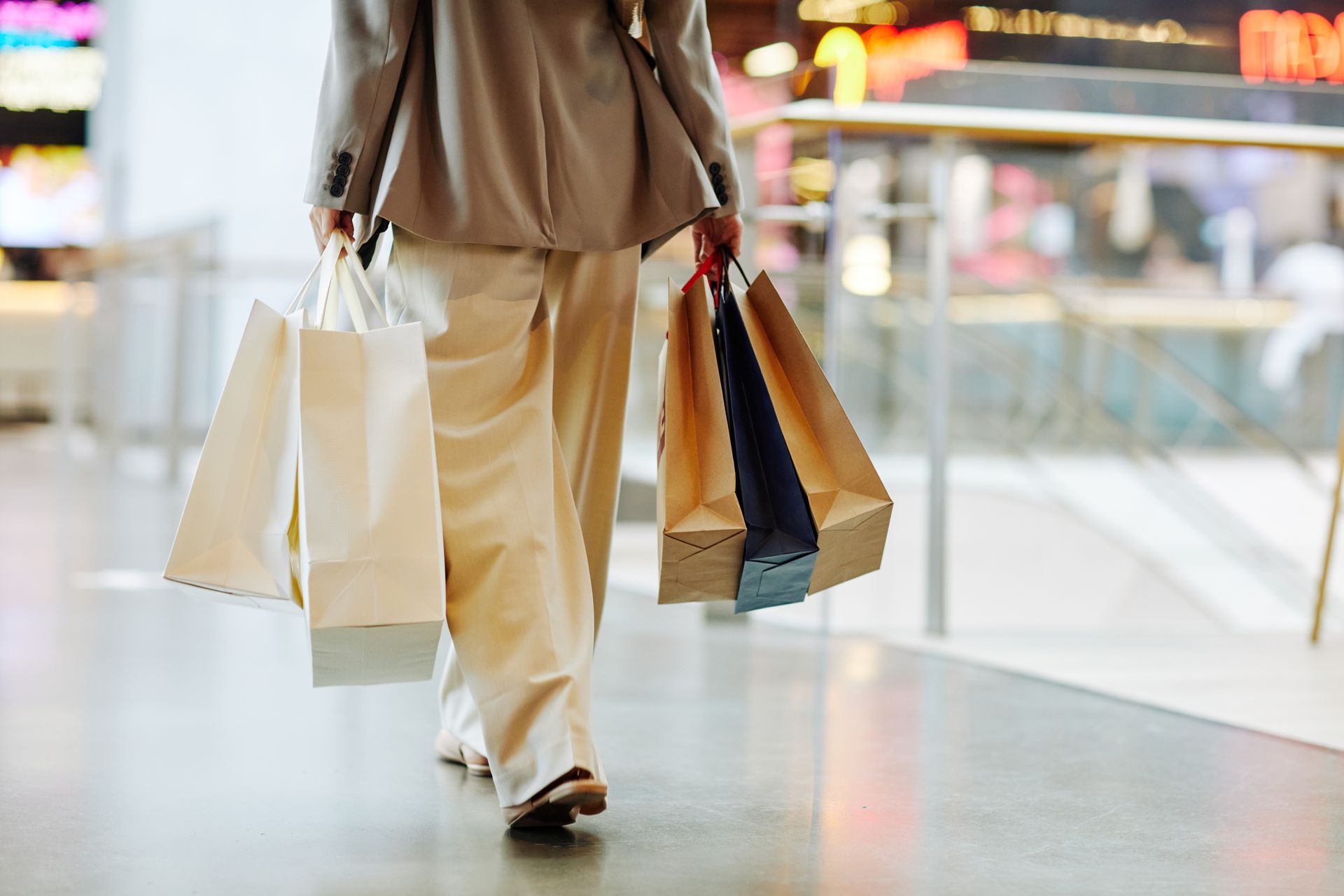 A low-angle, rear view of a person walking through a modern, brightly lit shopping mall corridor. 