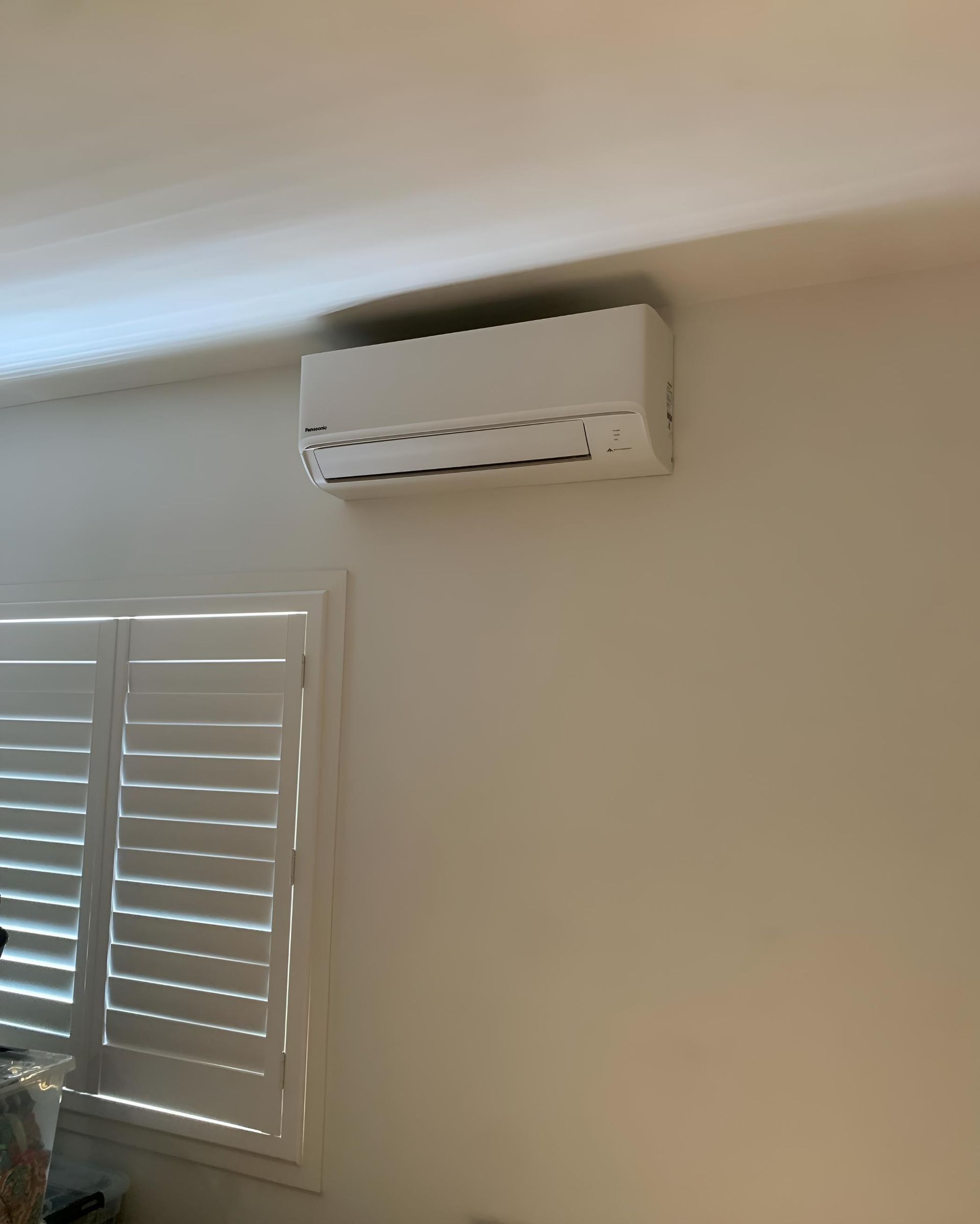 White air conditioner mounted on a beige wall, above a window with closed white shutters — Coolman Airconditioning in Broken Head, NSW