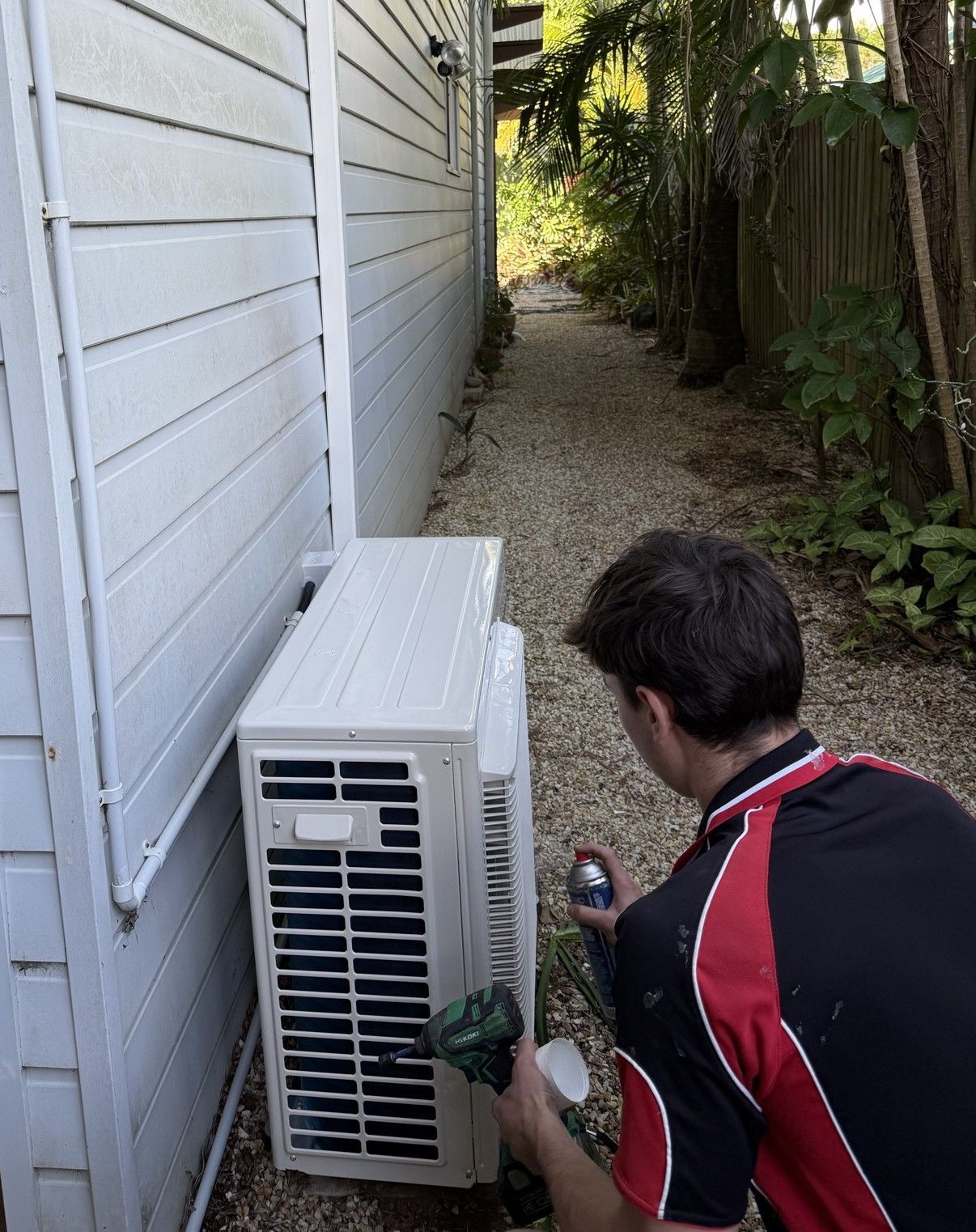 A person spraying an air conditioning unit outside a white building. Narrow pathway behind— Coolman Airconditioning in Goonellabah, NSW