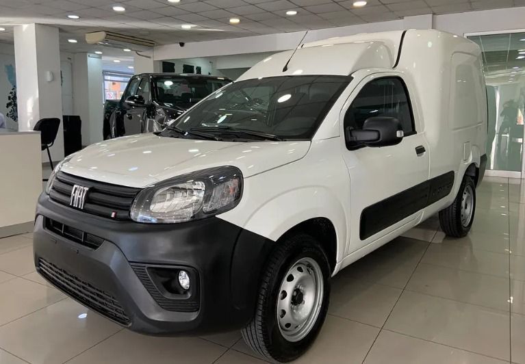 Furgoneta de carga Fiat Fiorino blanca en sala de exposición, parachoques delantero negro, ruedas grises.