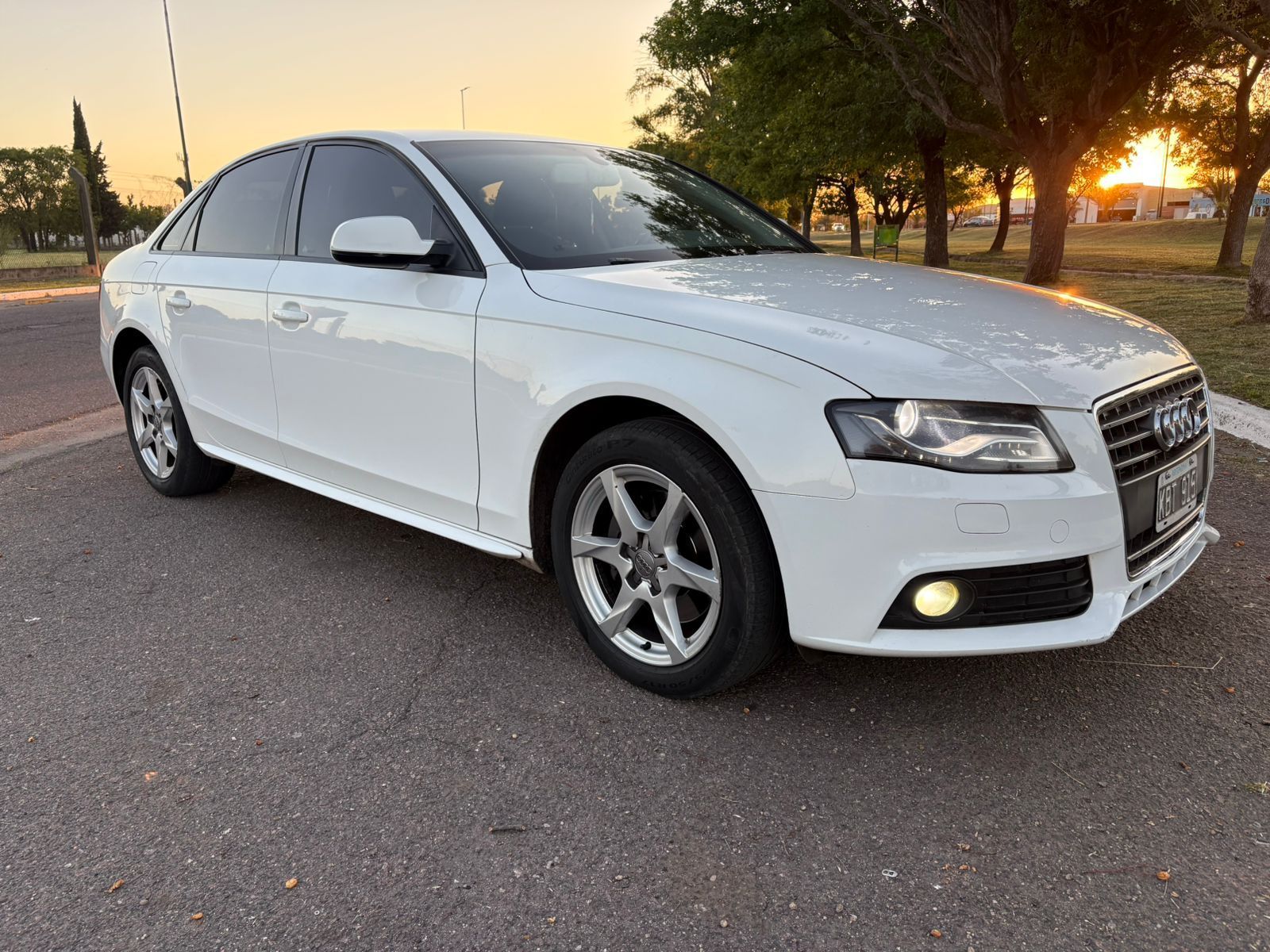 Audi A4 sedán blanco estacionado sobre asfalto, con árboles y puesta de sol al fondo.