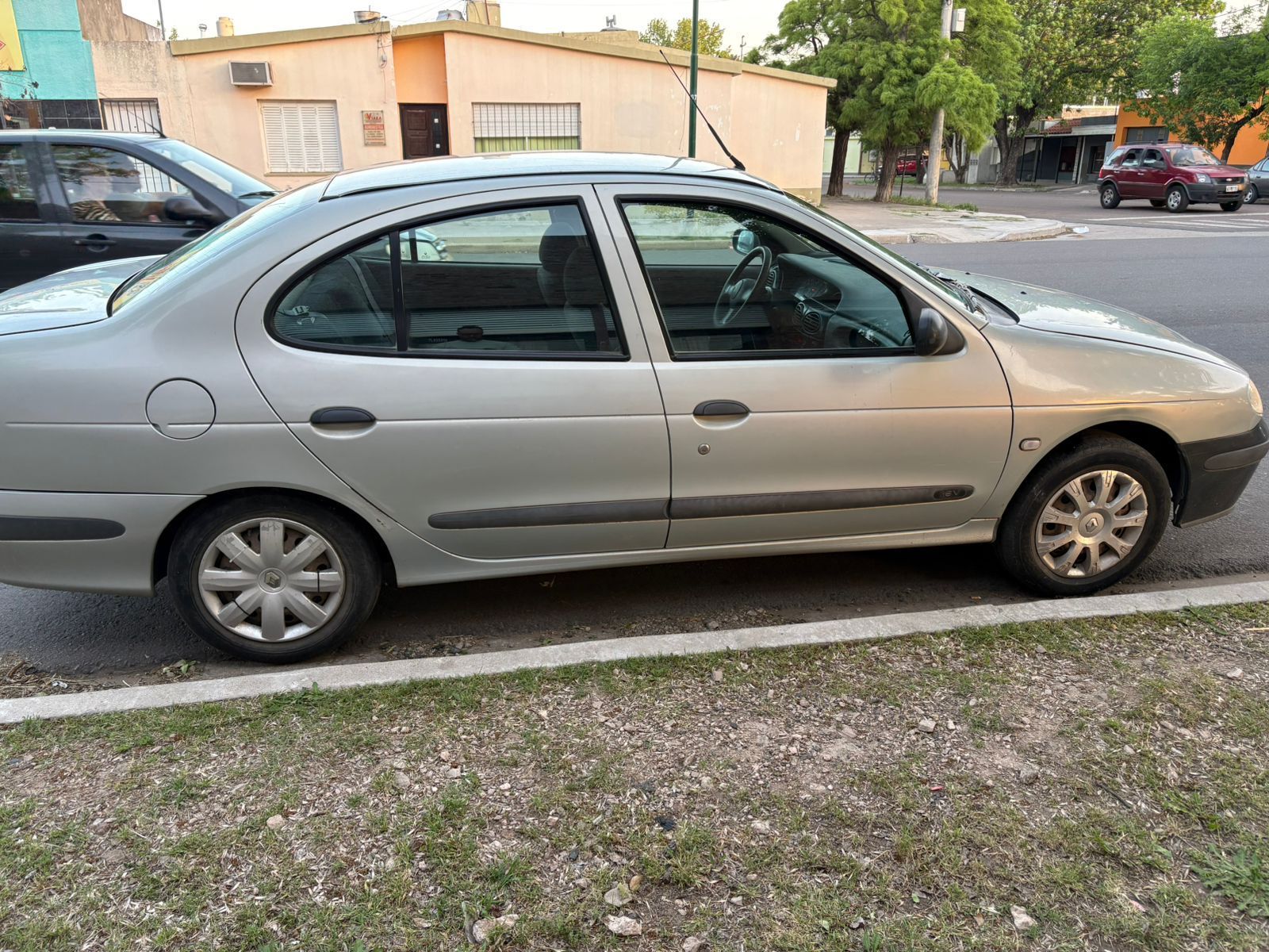 Renault Mégane sedán plateado estacionado al costado de una carretera, con edificios y una calle al fondo.
