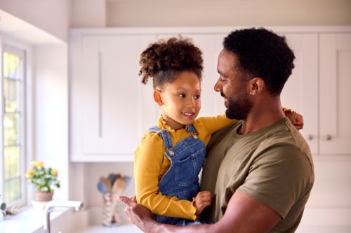 a black man wearing a navy green shirt carrying his female toddler wearing a yellow long sleeve shirt under a denim jumper in the kitchen