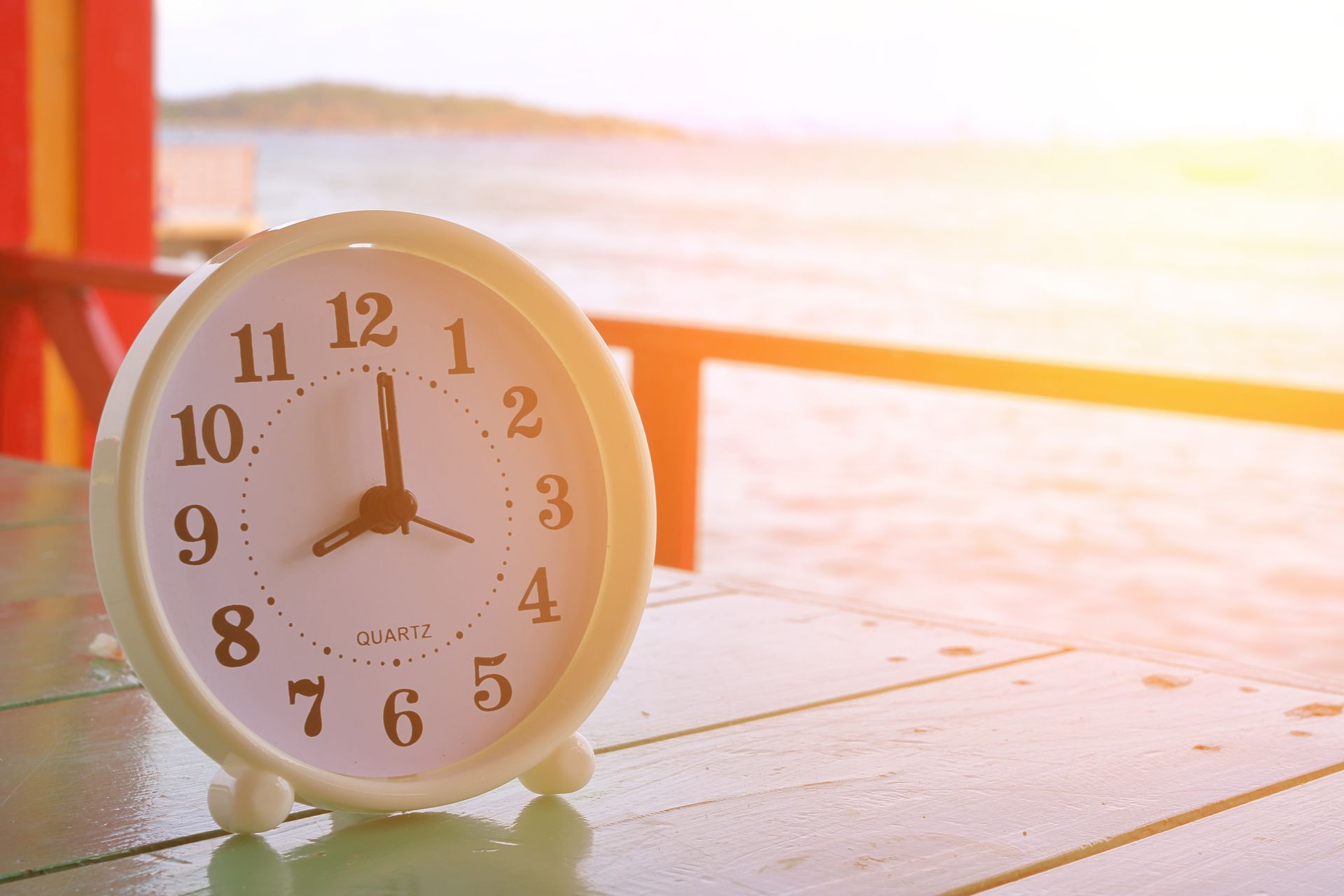 image of an analog desk clock showing 8AM on a wooden table by the beach.