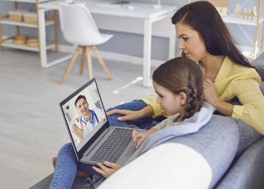 A mother sitting beside her young daughter on a couch at home while on a video call with a healthcare professional virtually on a laptop