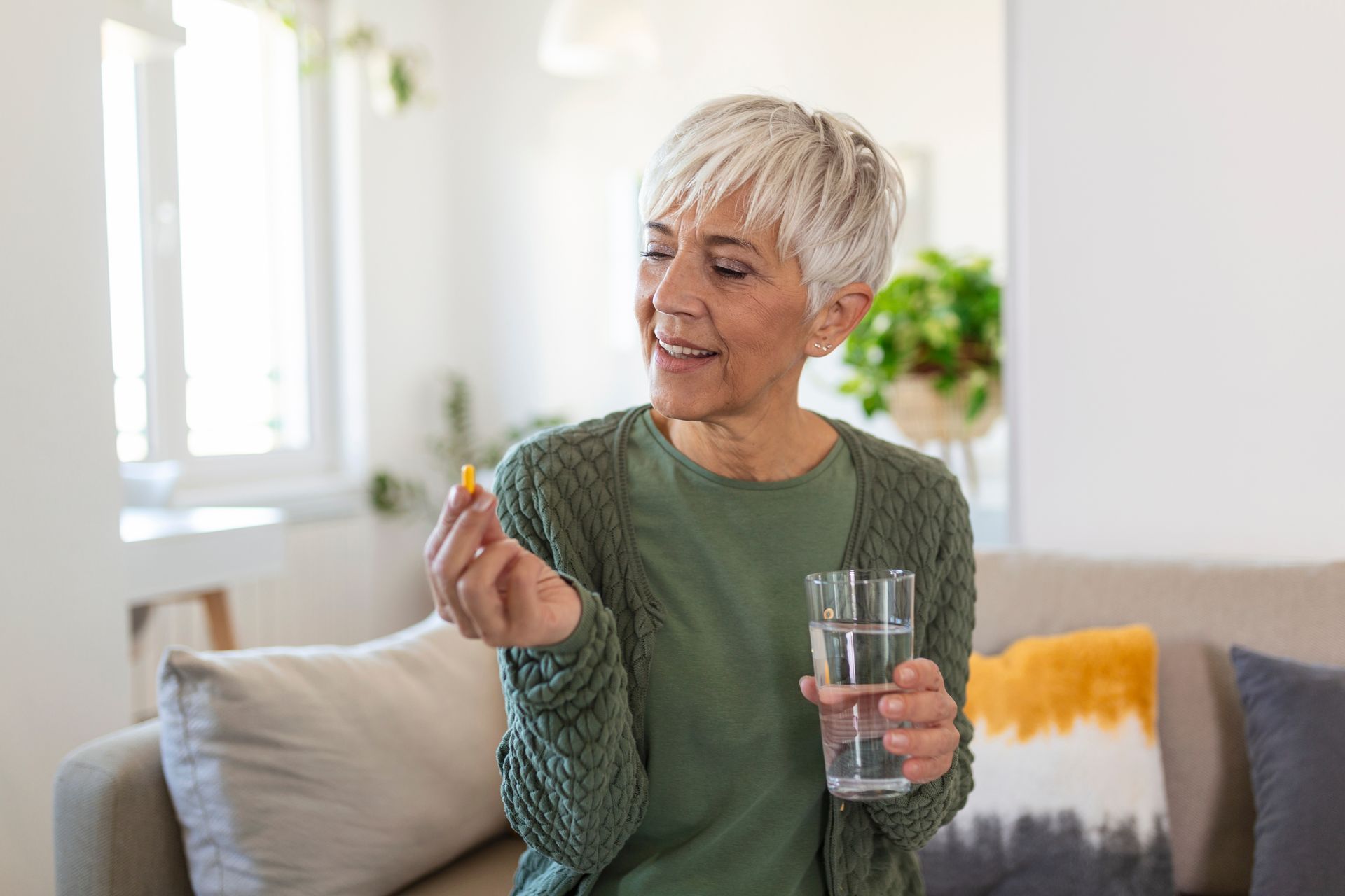 image on a elderly woman wearing a green cardigan and shirt. Holding a pill on one hand and a glass of water on the other.