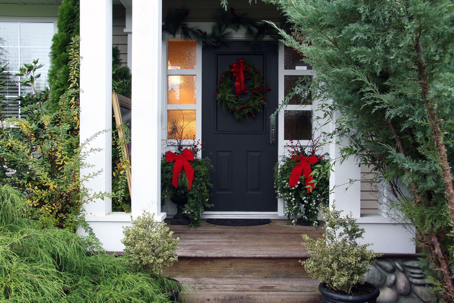 A black front door decorated with wreaths and red bows, flanked by white columns and greenery, Christmas scene.
