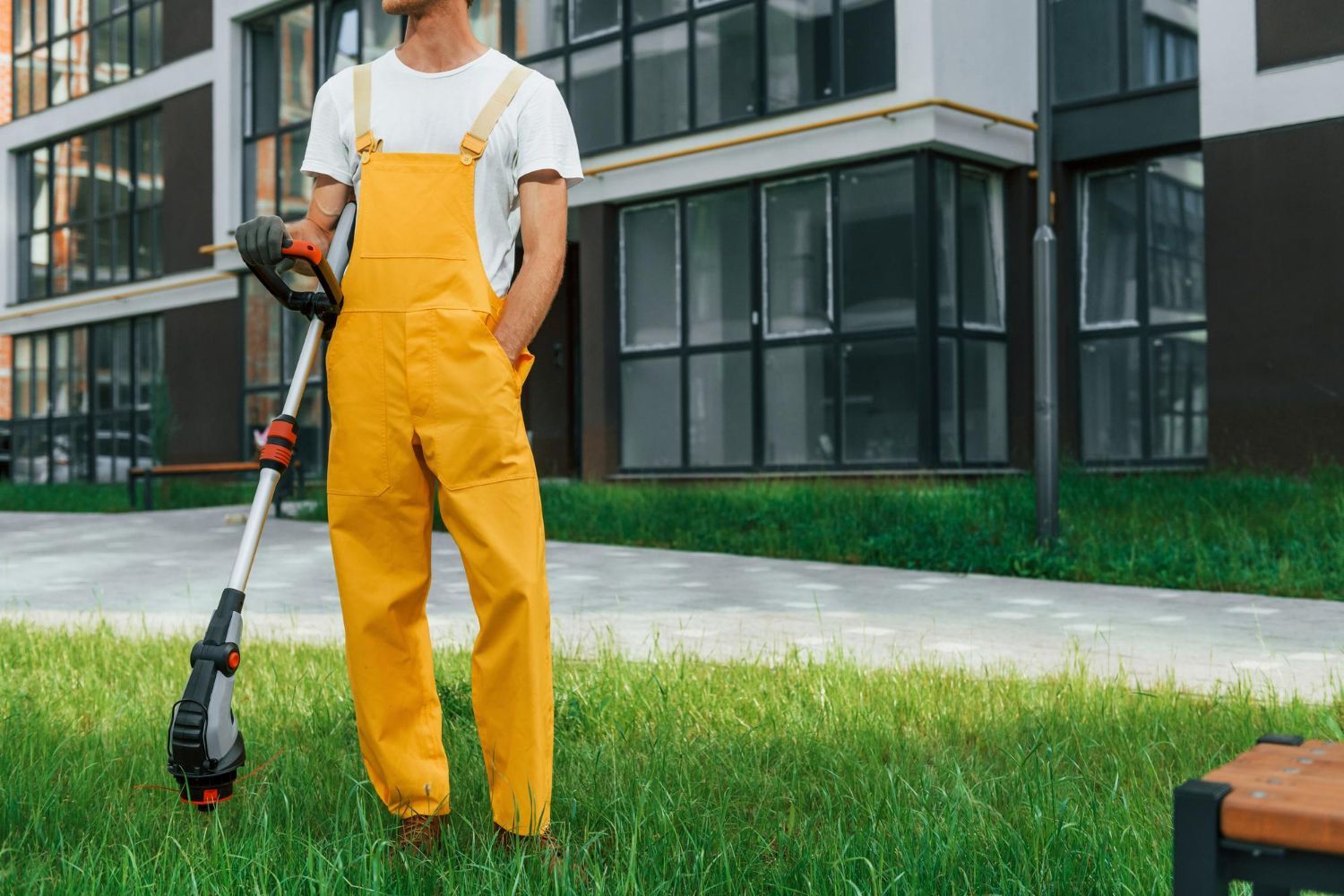 A man in yellow overalls is standing in the grass holding a lawn mower.