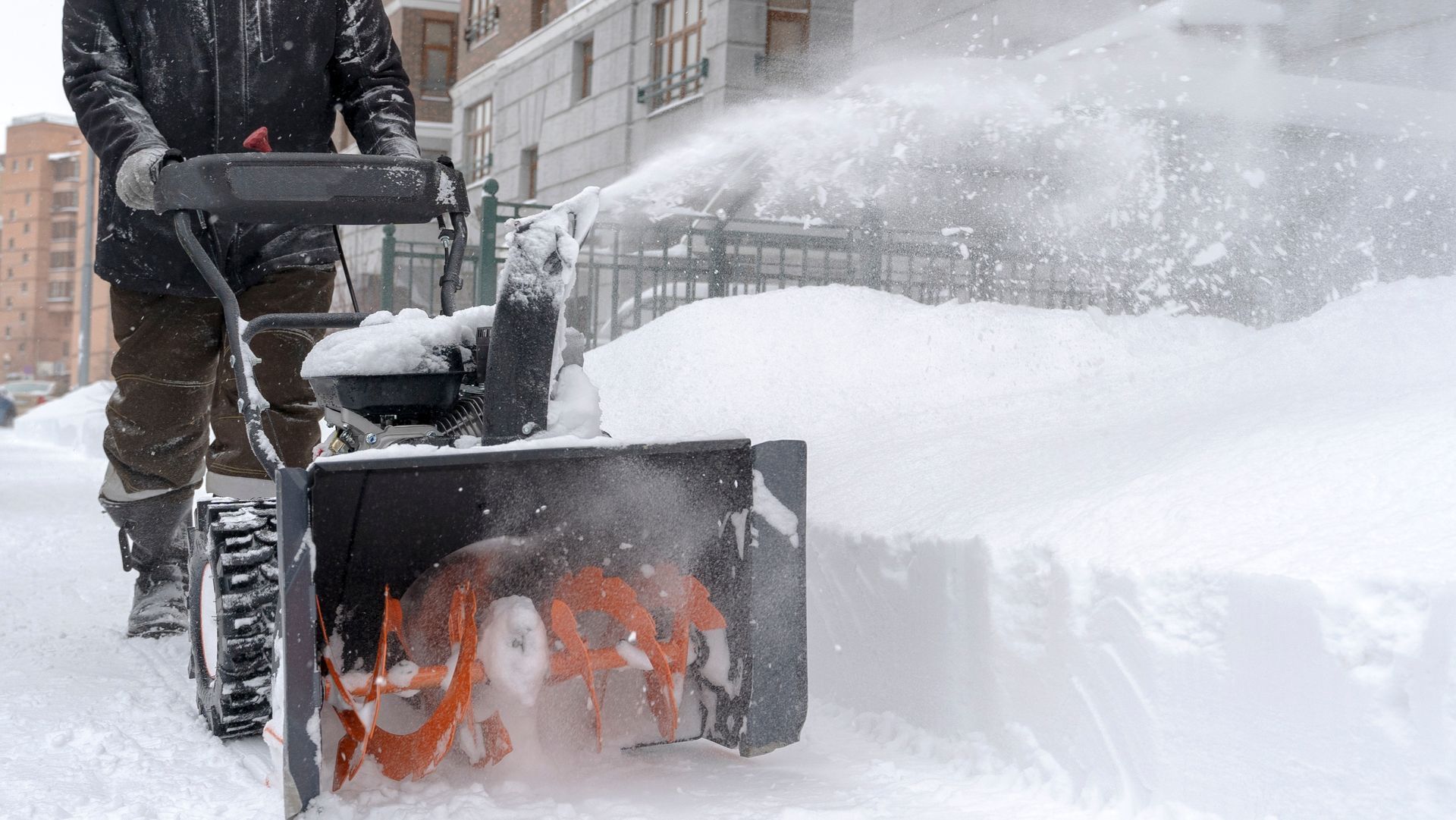 Person using a snow blower to clear a sidewalk covered in deep snow.