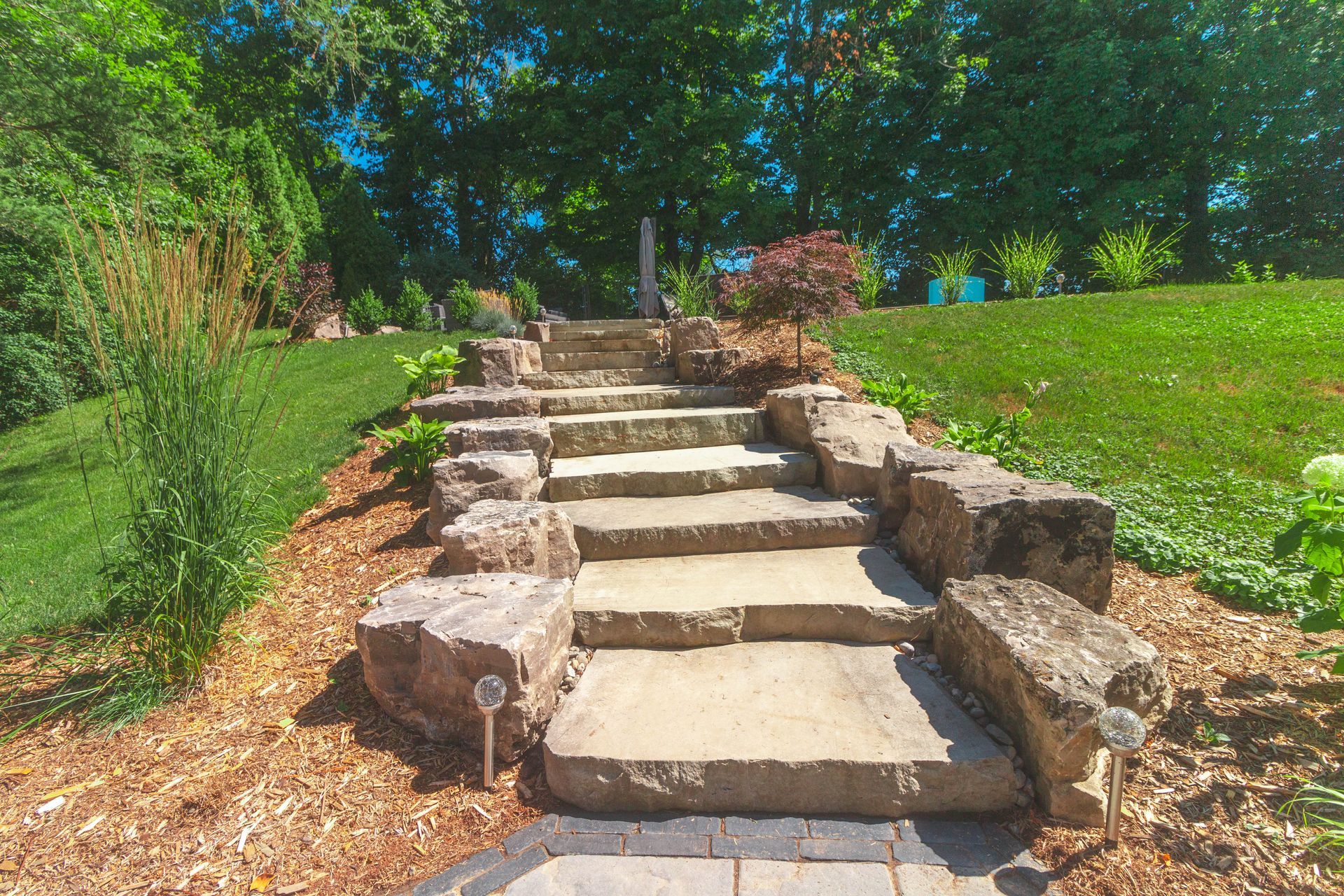 Stone steps lead up a grassy hill, edged with rocks and mulch, trees in background.