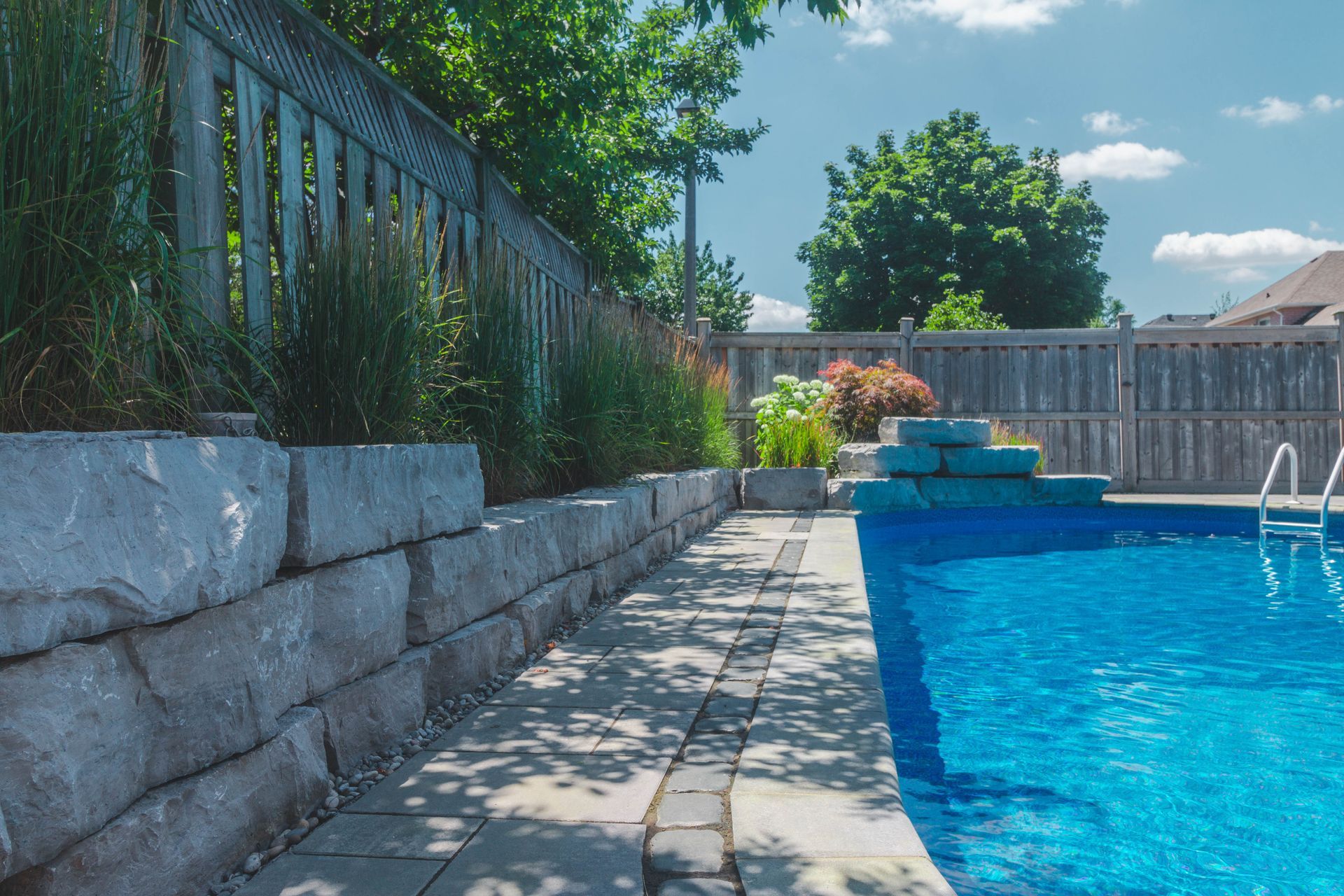 Stone retaining wall with plants next to a pool. Blue water, wooden fence, sunny day.