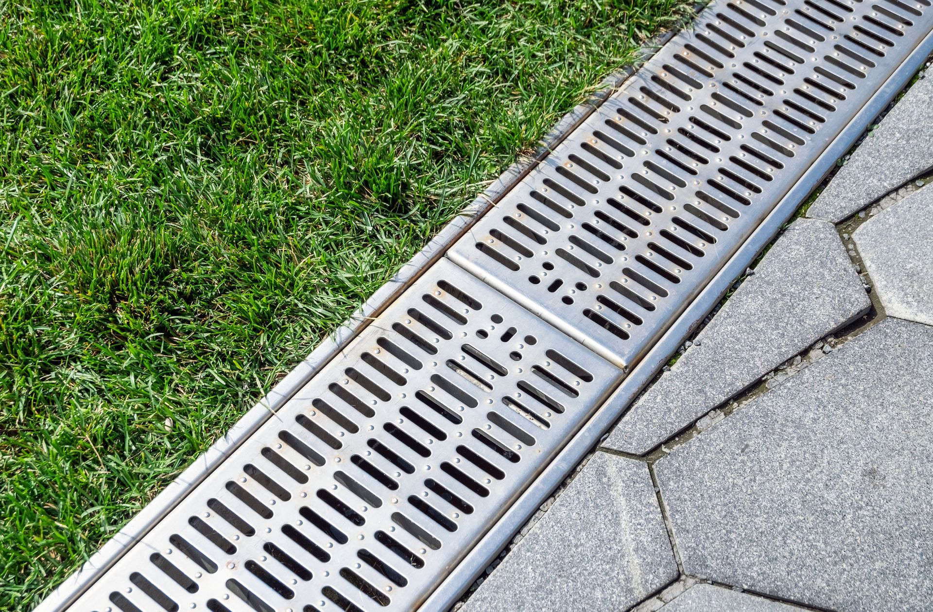 Metal grate drain on gray stone paving next to green grass.