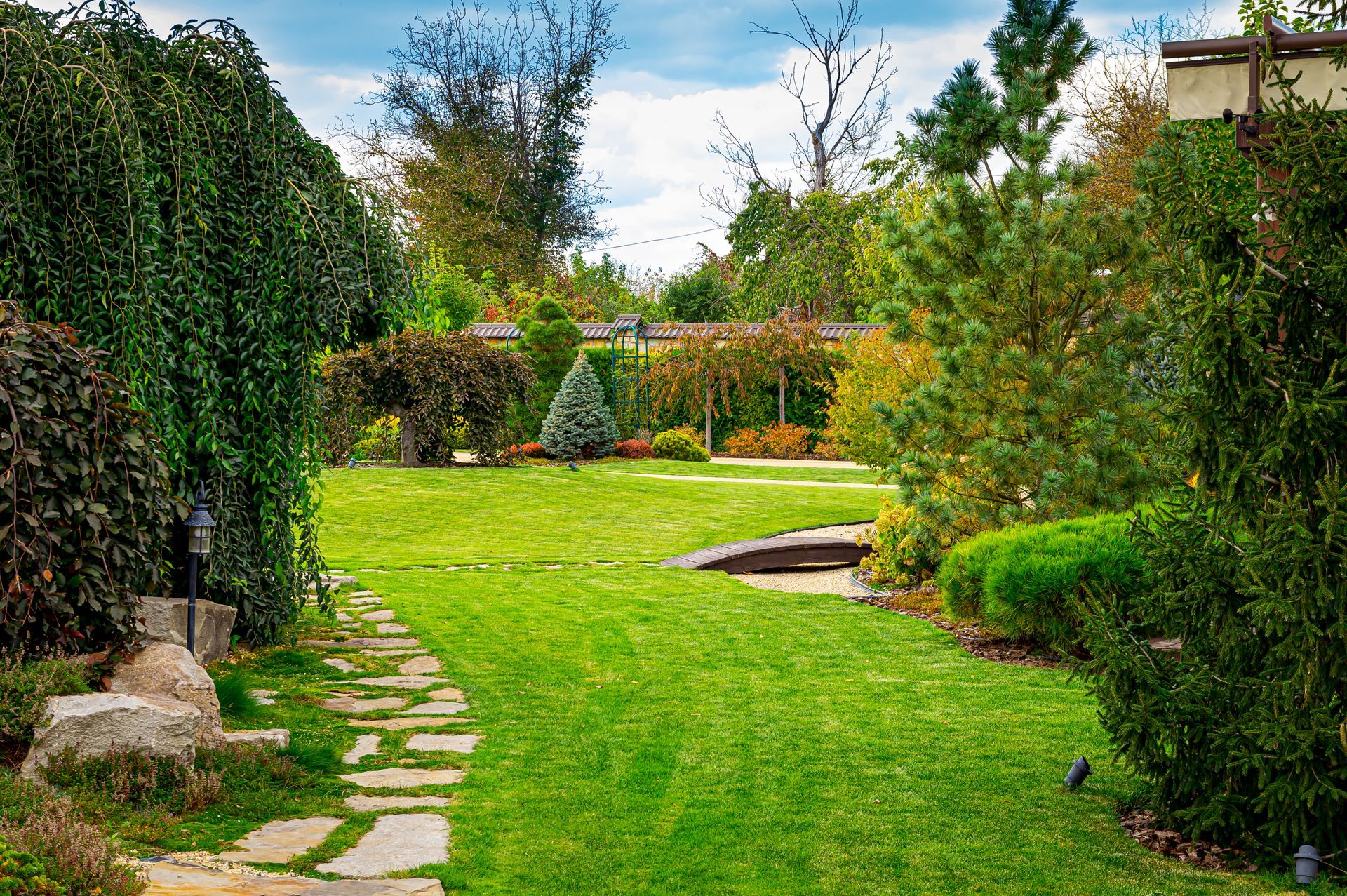 A stone path curves through a lush green lawn in a landscaped garden with trees, shrubs, and a distant trellis.