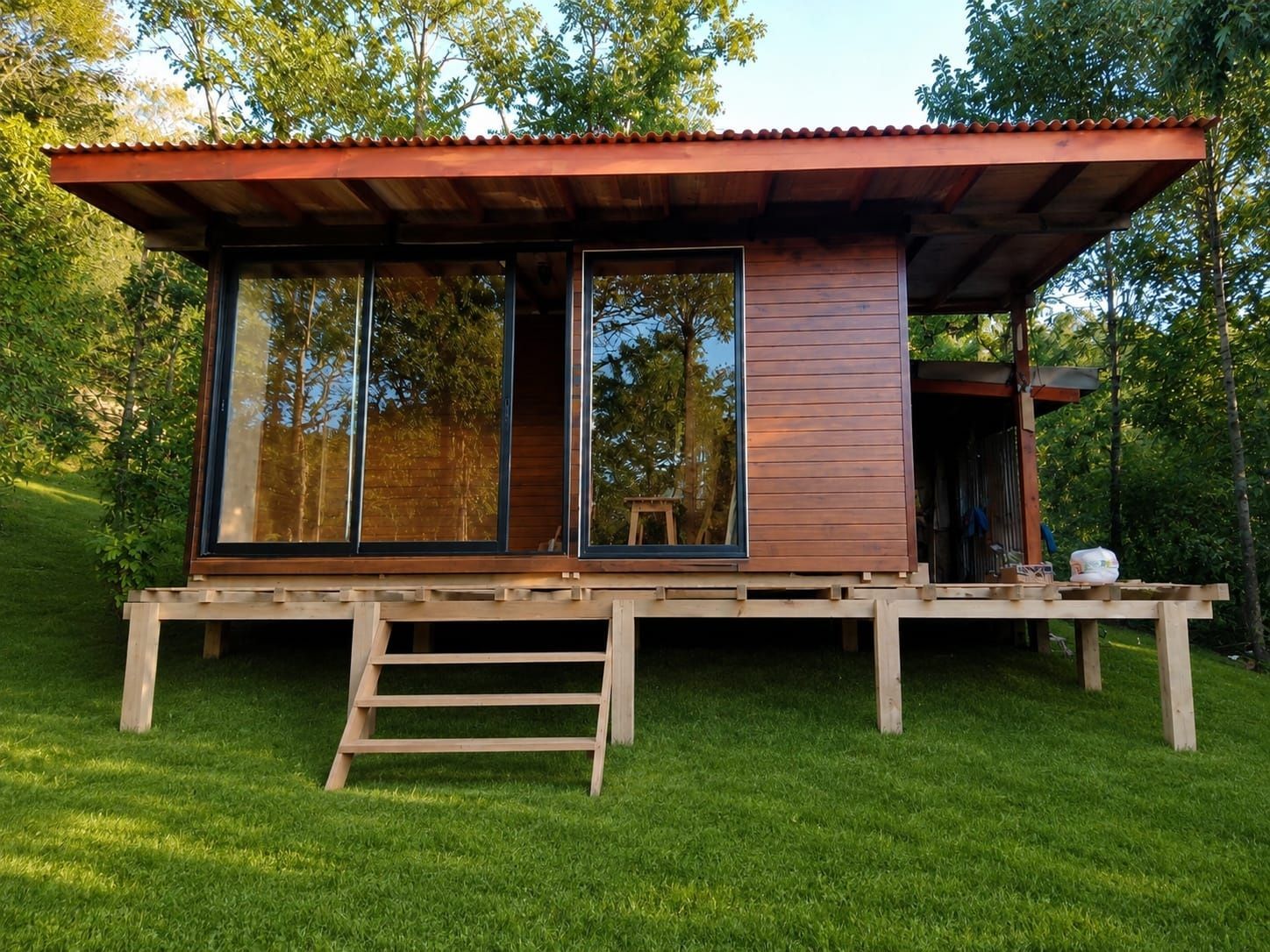 Pequeña cabaña moderna de madera con grandes ventanales y techo rojo en un entorno boscoso y cubierto de hierba.
