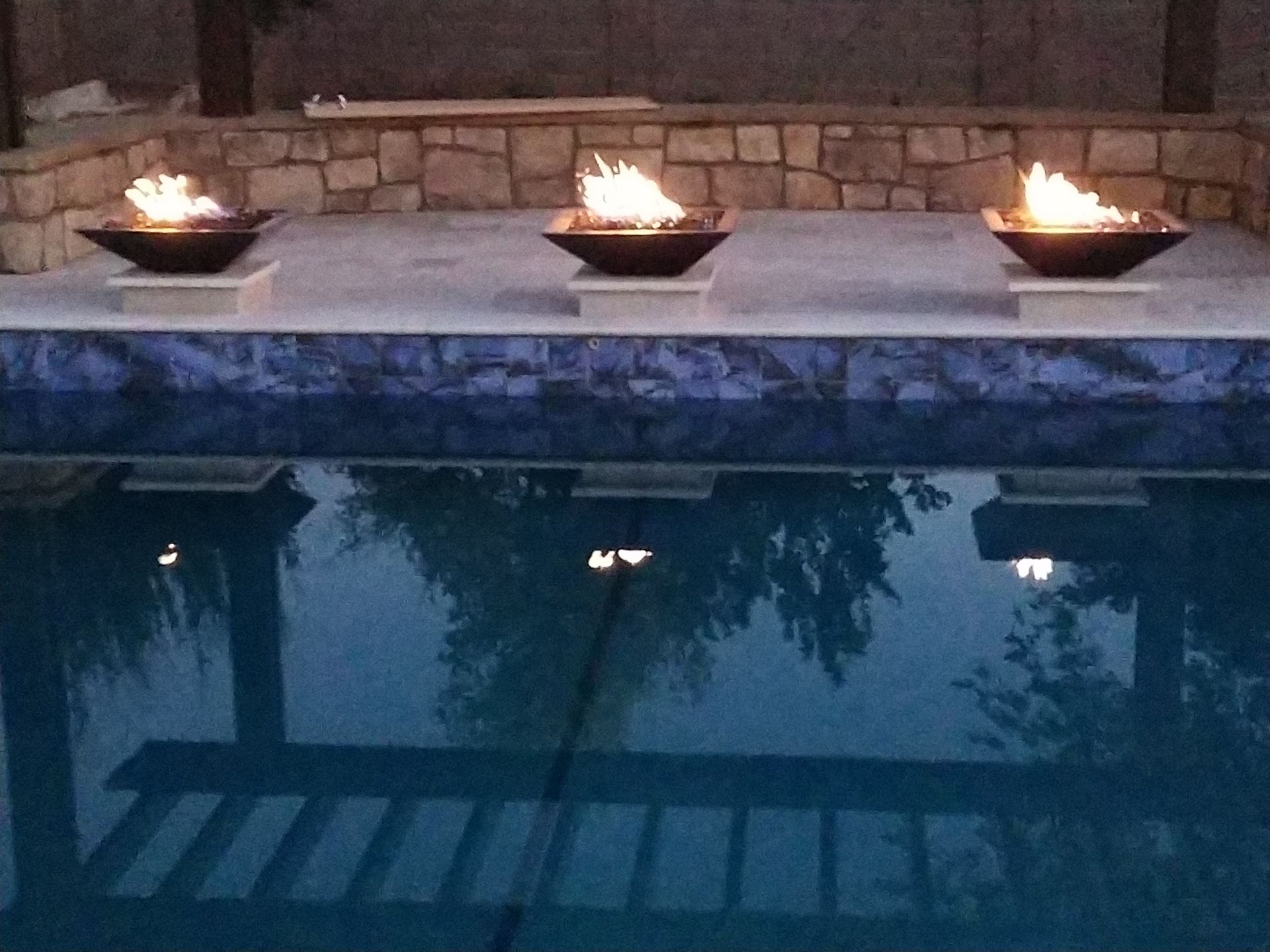 Three large, light gray planters with green plants sit on a brick walkway in front of a building with brick and windows.