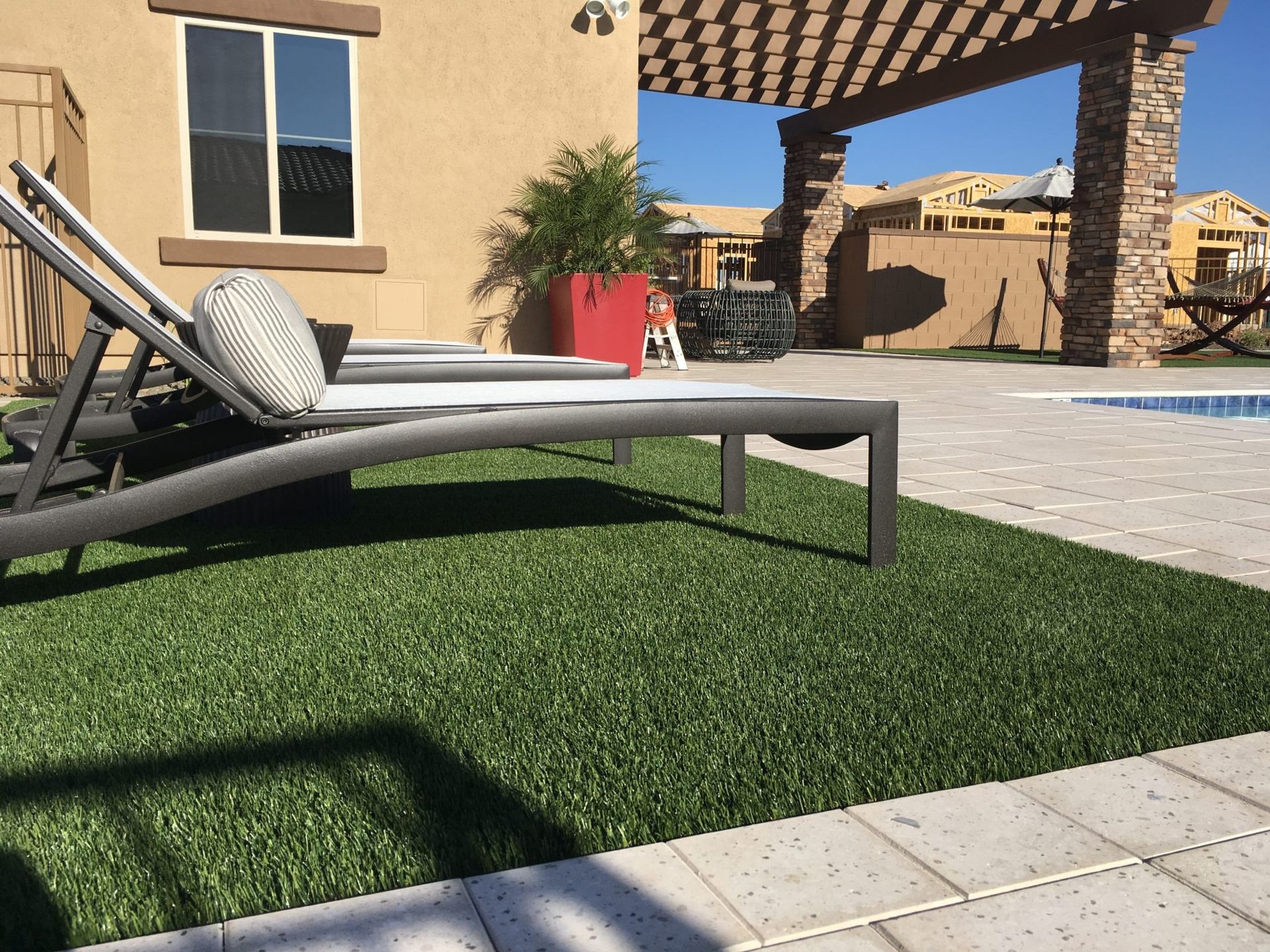 Lounge chairs on green turf beside a pool with a pergola and stucco home. Sunny outdoor scene.