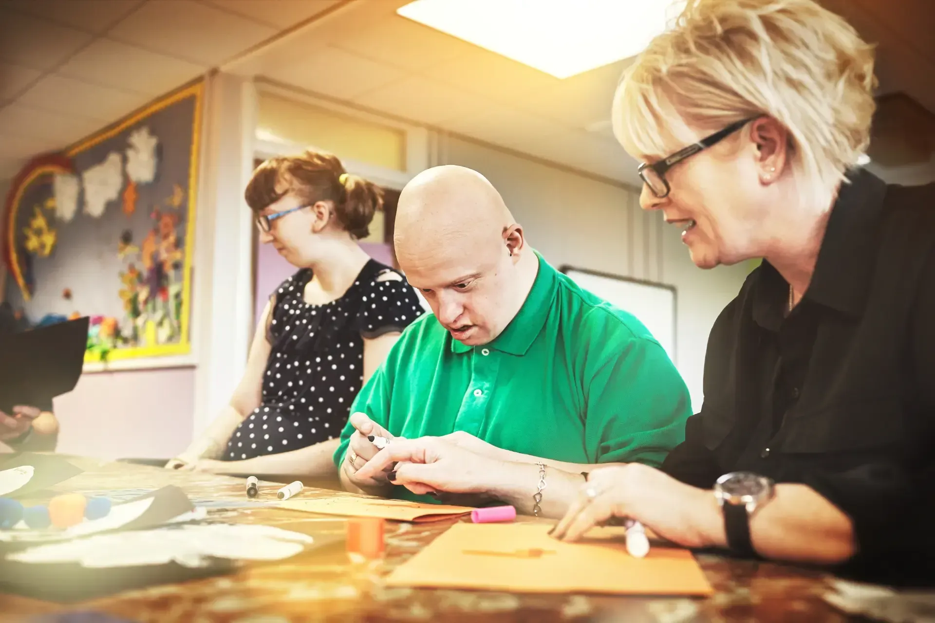 Woman pointing upwards with a young person, possibly with Down syndrome, sitting close. They are at a table with toys.