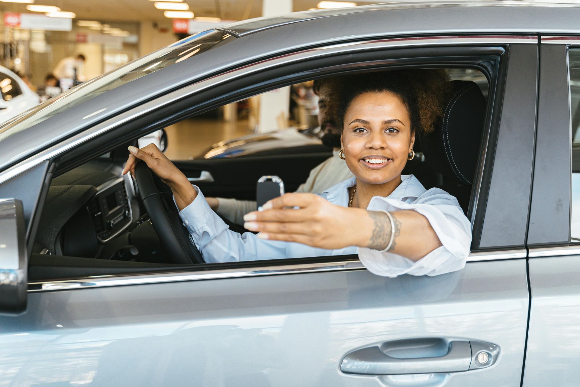 Woman in a silver car smiles, holding out car keys. She is inside a car dealership.