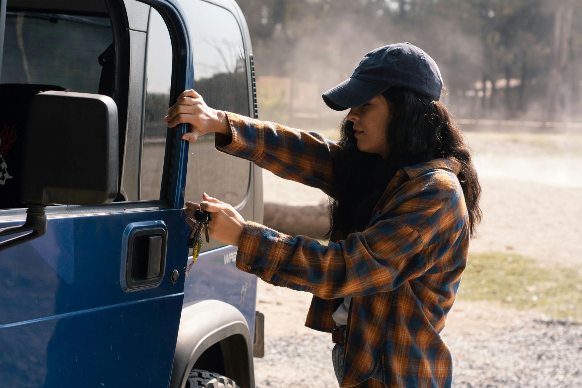 Woman in a flannel shirt and cap opening the door of a blue Jeep, outdoors on a sunny day.