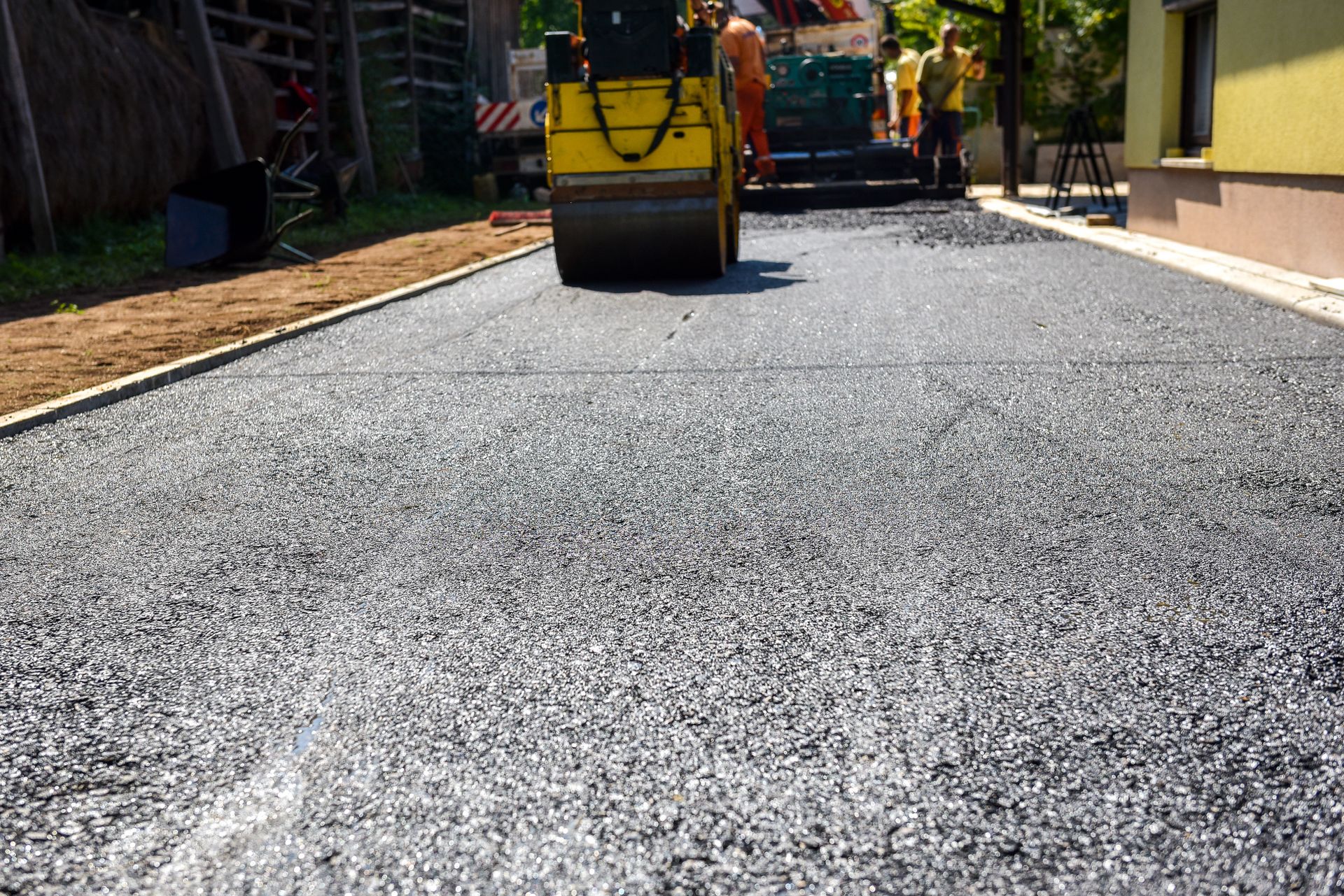 A yellow industrial road roller compacts freshly laid dark asphalt on a sunny outdoor construction site.