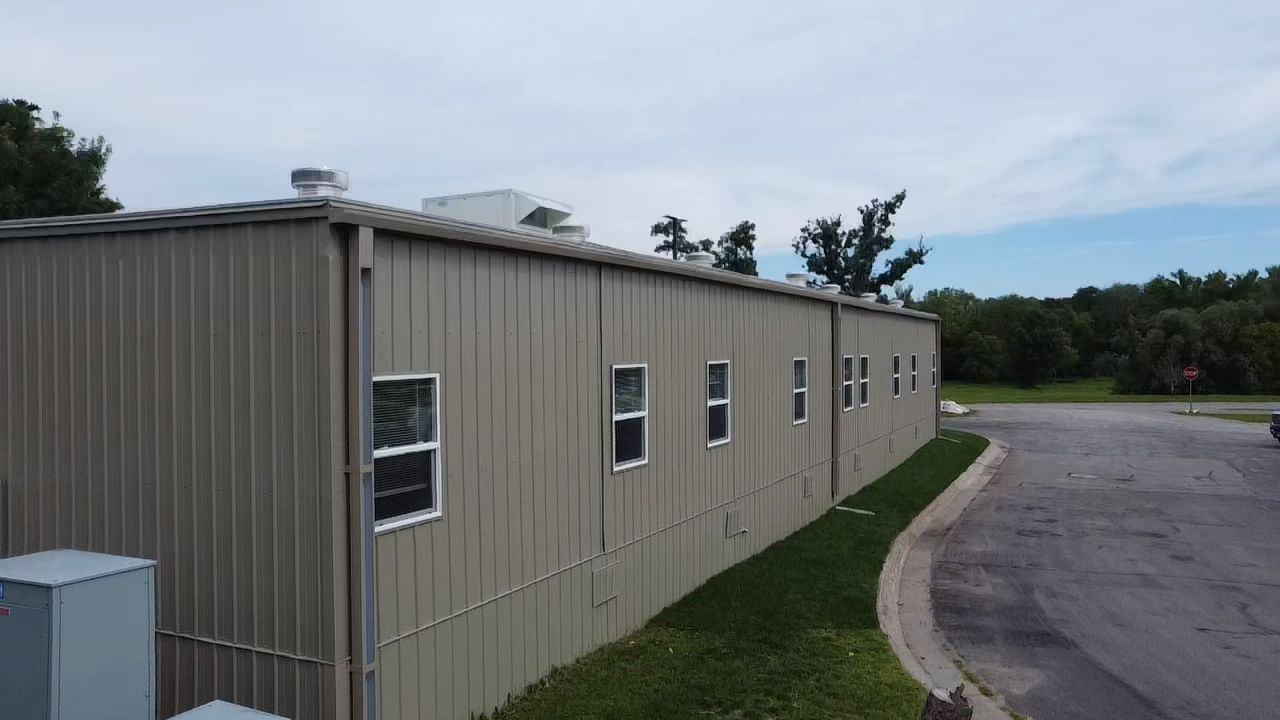 Tan metal building with multiple windows, next to a curving road and green lawn. Overcast sky.