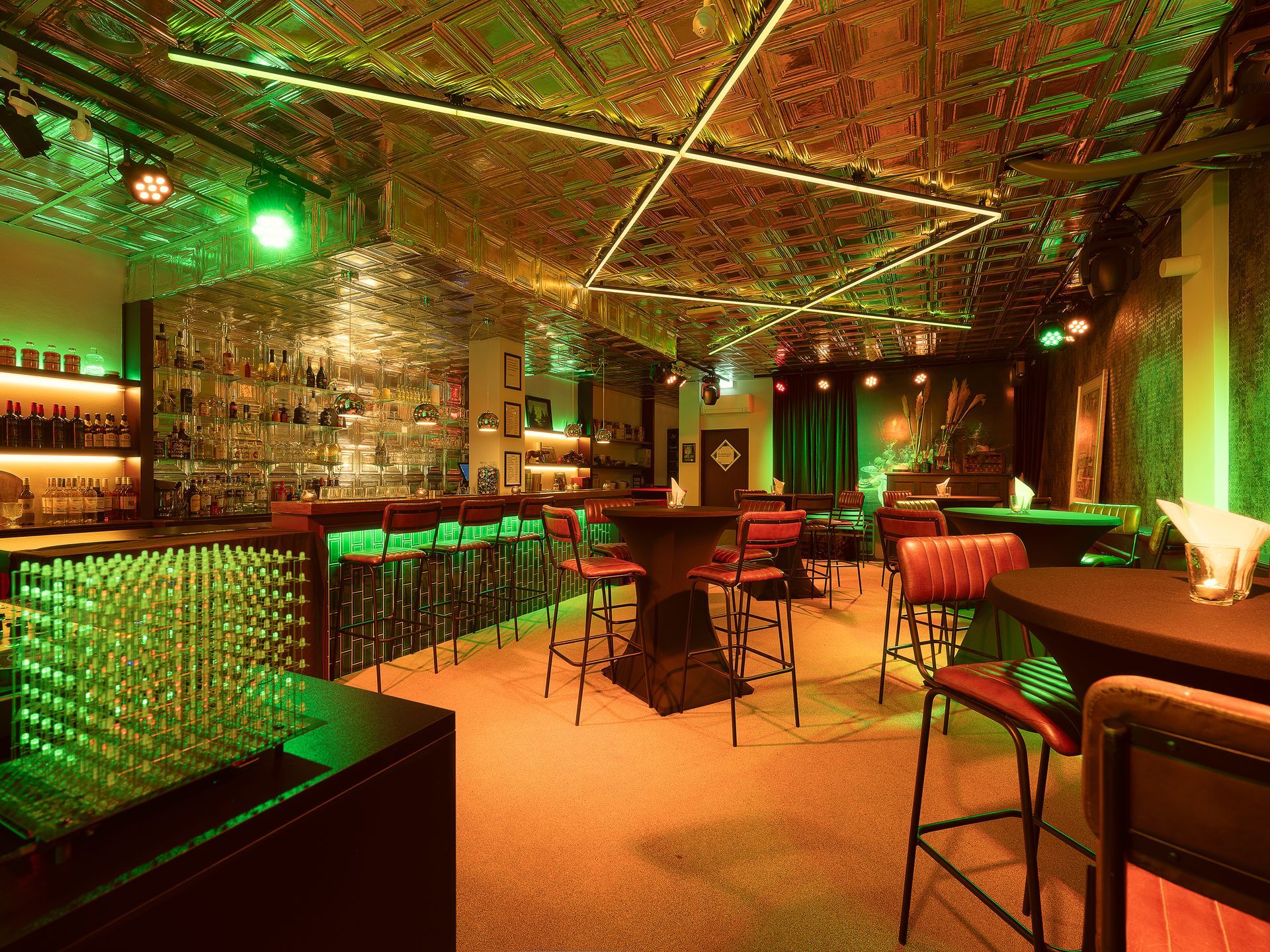 Interior of a bar with green and gold lighting, a long bar, and red bar stools.