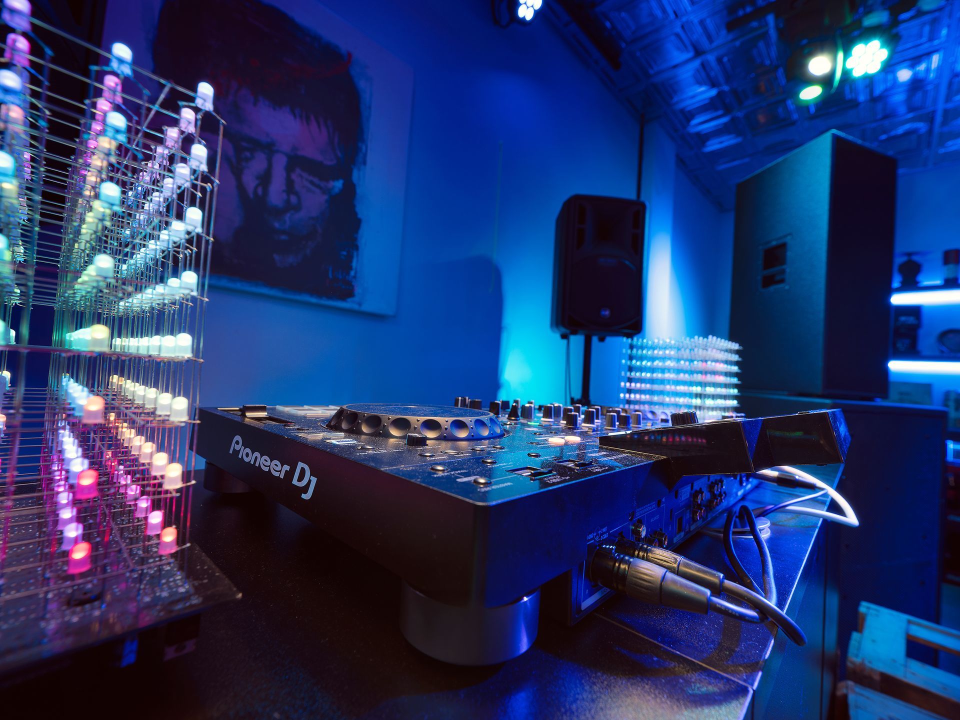 DJ setup: Pioneer DJ mixer, speakers, LED light display in a blue-lit room with art.