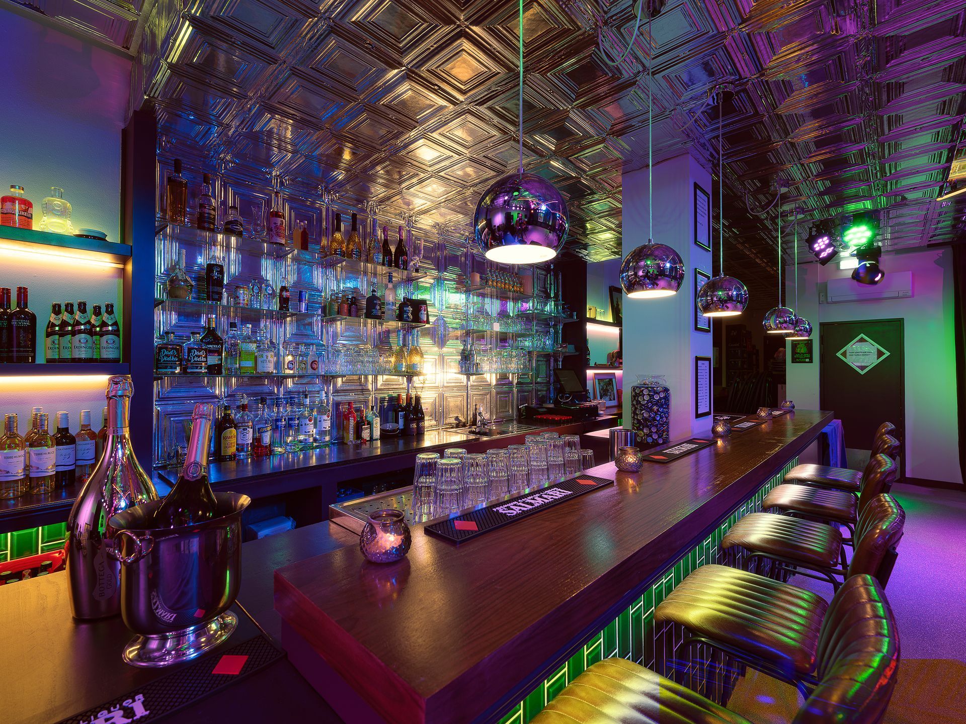 Bar interior with shiny ceiling and bottles, glasses, and chairs at the counter, lit with colorful lights.