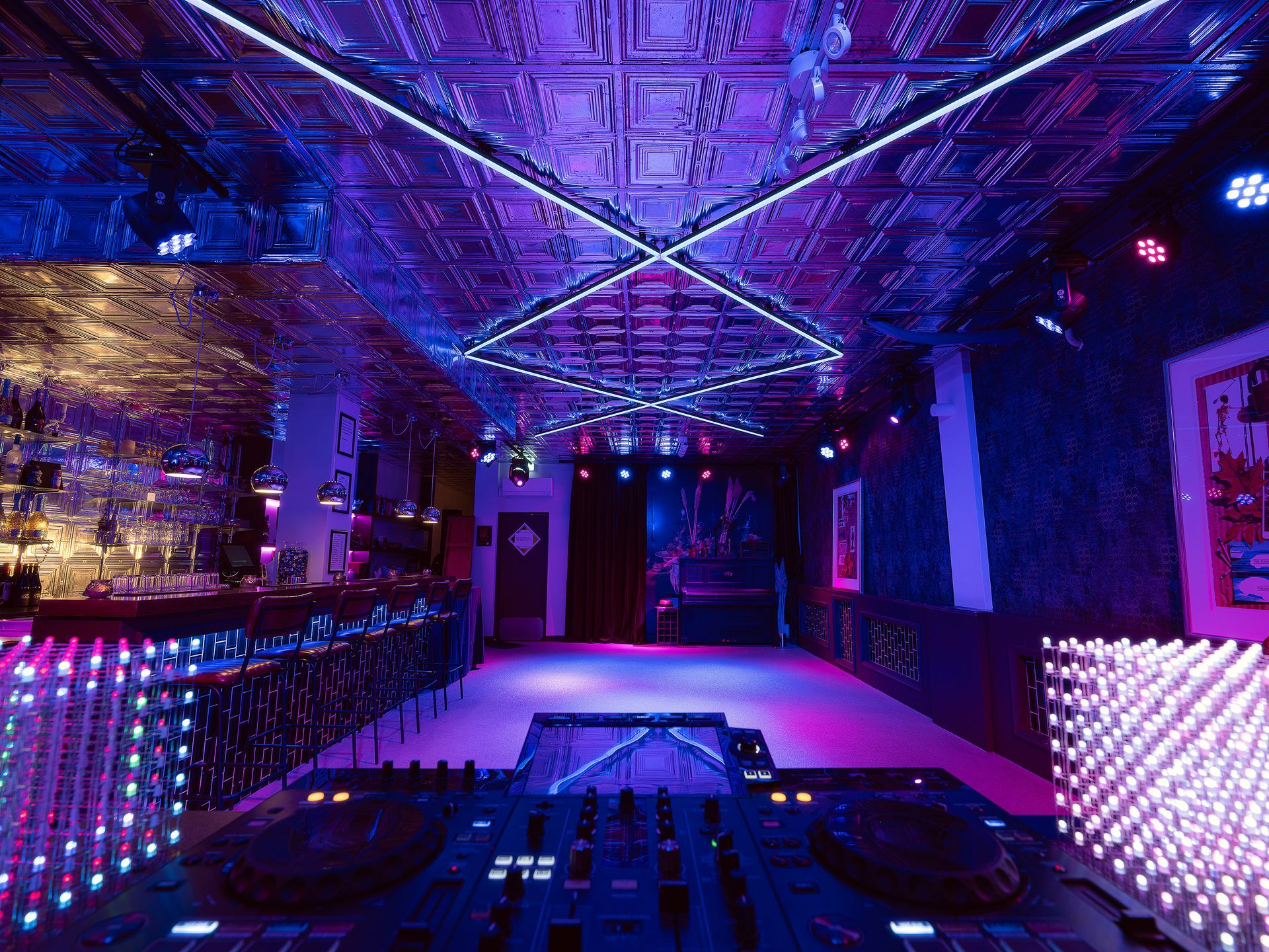 DJ setup in a dimly lit club. Purple and blue lights illuminate a dance floor and bar area.