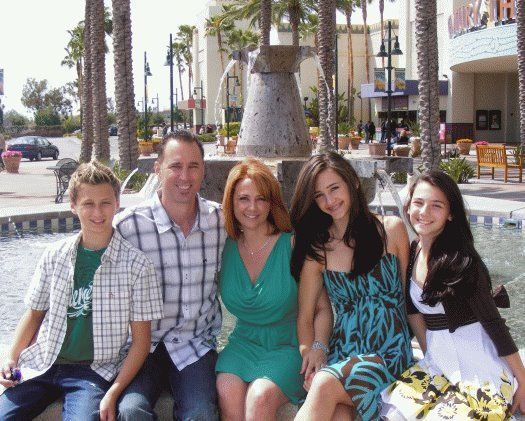 Family smiling, posing by fountain. Boy in plaid shirt, girls in dresses, sunny day, palm trees.