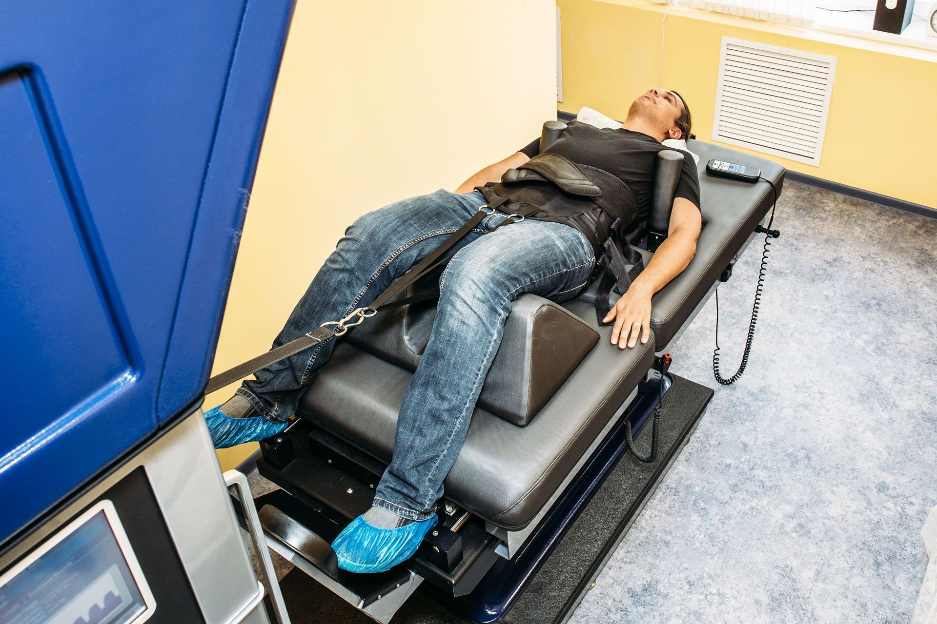 Person lying on treatment table, wearing straps. Blue machine to left. Bright room.