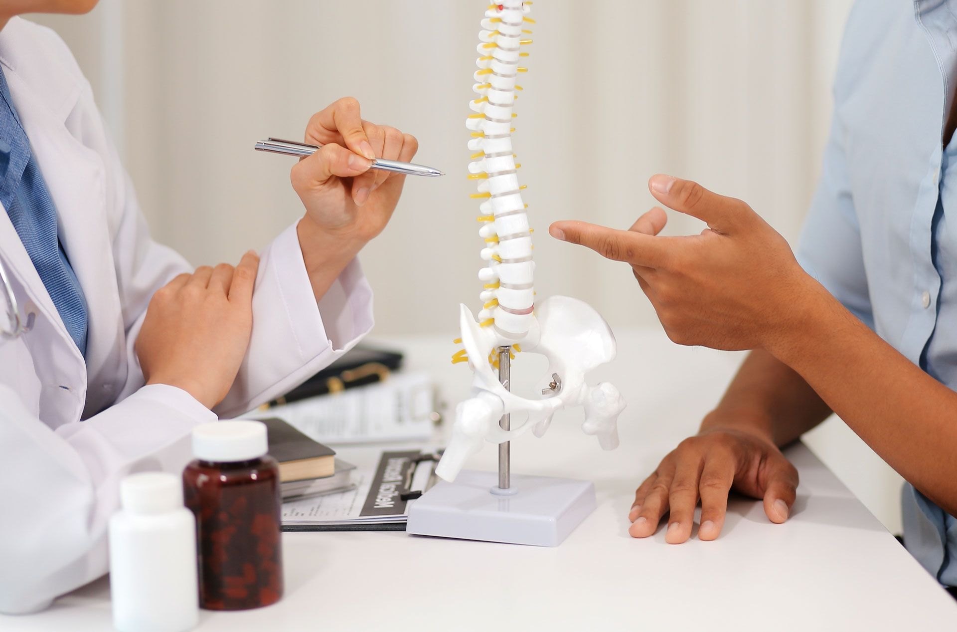 Doctor pointing to spine model, explaining to patient. Desk with medication bottles.