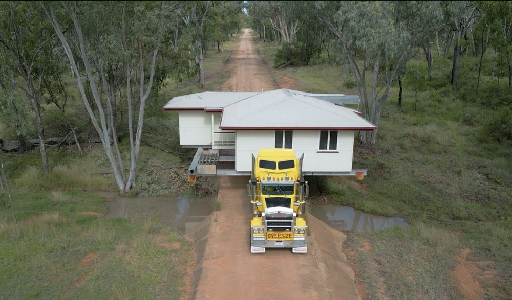 A Yellow Semi-truck Transports a White House Down a Dirt Road Lined With Trees — Pro-Jax Pty Ltd in Bohle, QLD