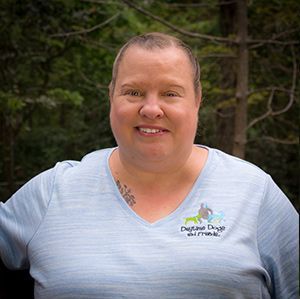 Susan in a gray shirt is smiling in front of a forest.
