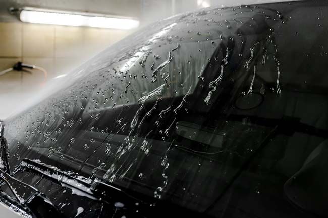 Black car windshield covered in water droplets, being washed under bright lights