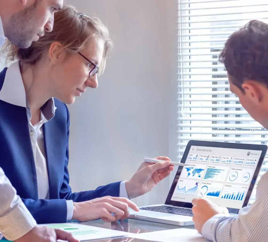 Three people at a table, looking at laptop screen with business graphs, discussing data.