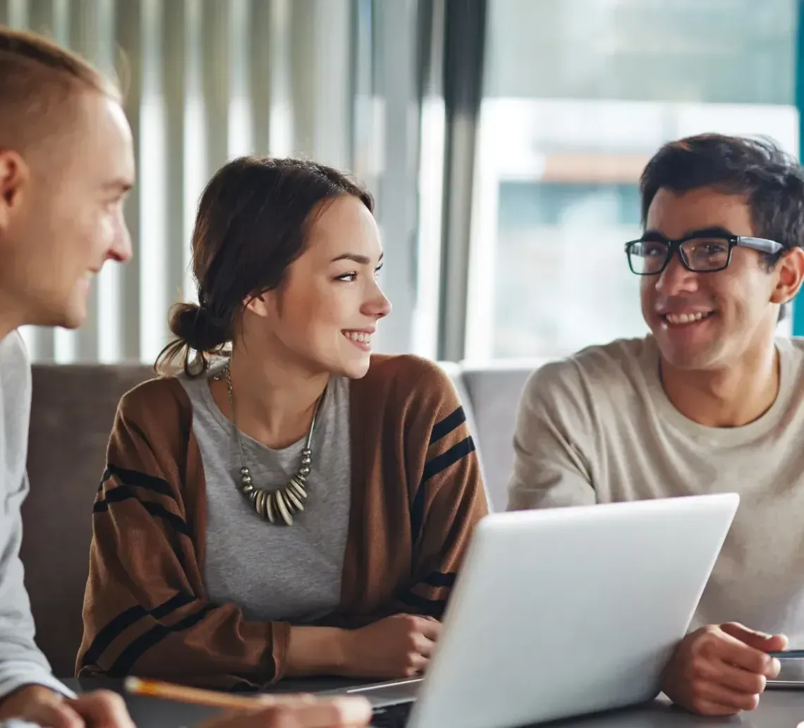Three people at a table with a laptop, smiling and talking. Bright, casual setting.
