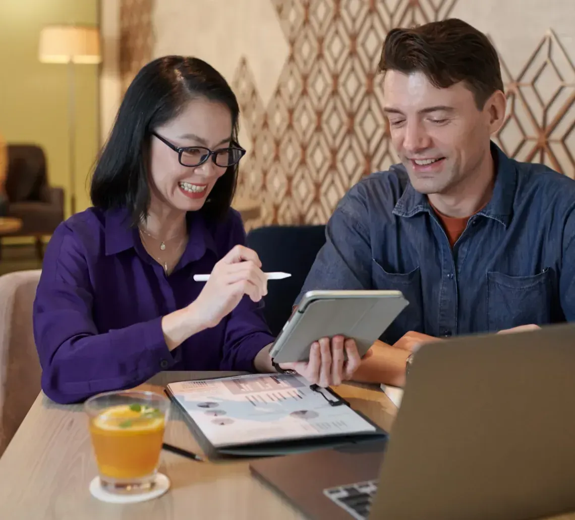 Two people looking at a tablet and laptop at a table, smiling. Drinks nearby, in a modern interior.