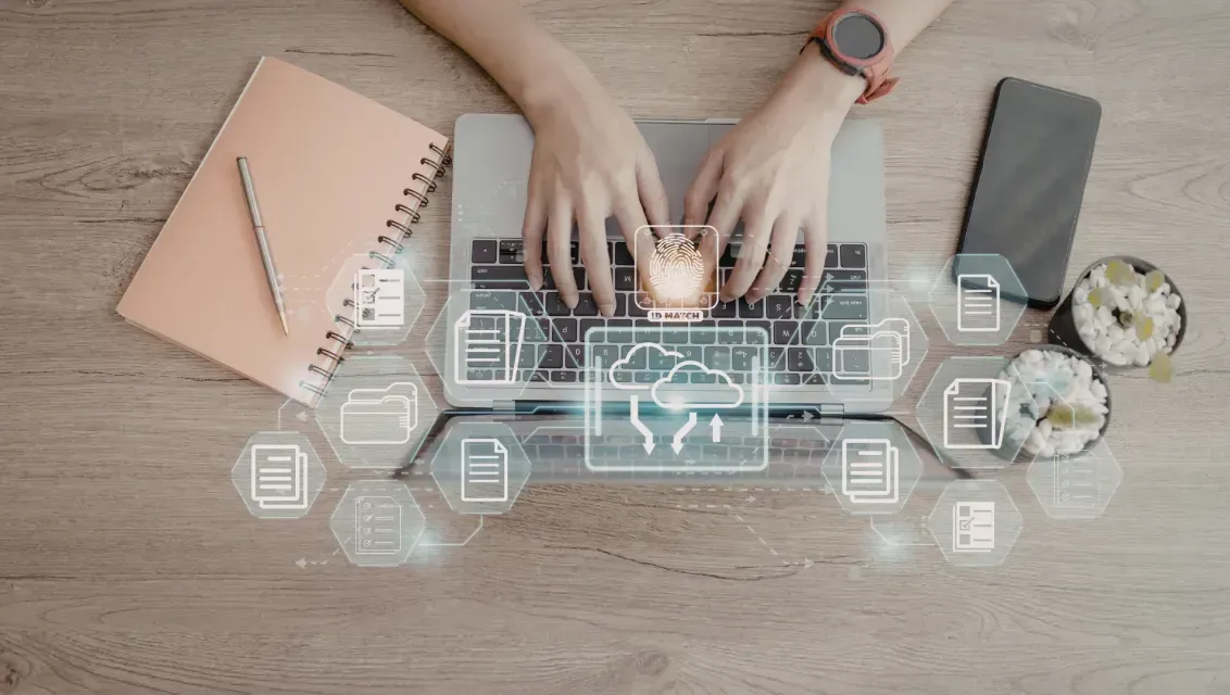 Hands typing on a laptop with cloud data icons overlaid on a wooden desk with a notebook, pen, phone, and plant.
