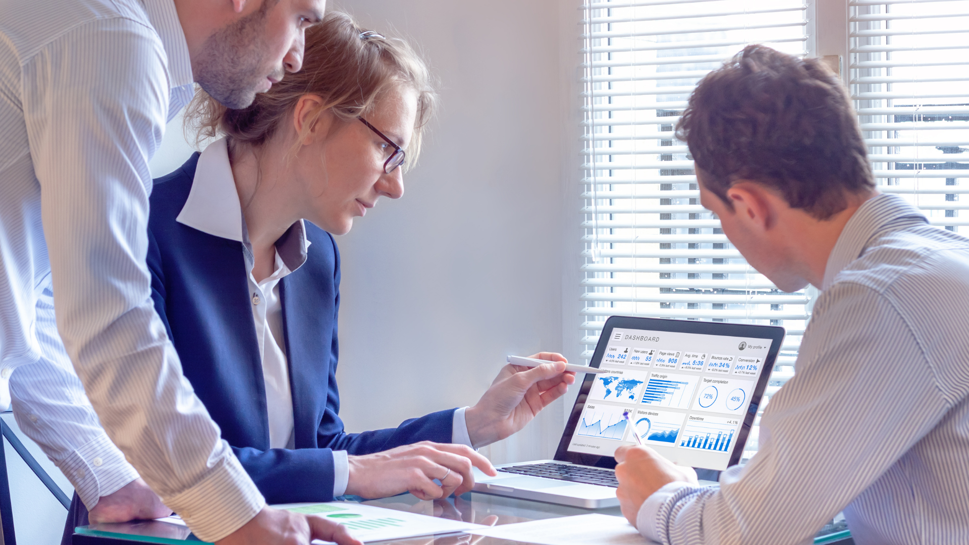 Three people at a table, looking at laptop screen with business graphs, discussing data.
