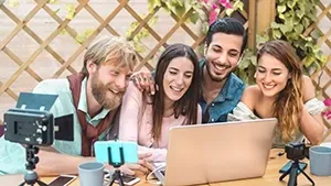 Four people laugh while looking at a laptop and small cameras, outdoors.
