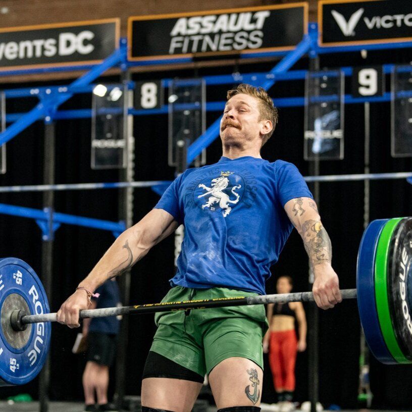 A man in a blue shirt is lifting a barbell at an assault fitness event