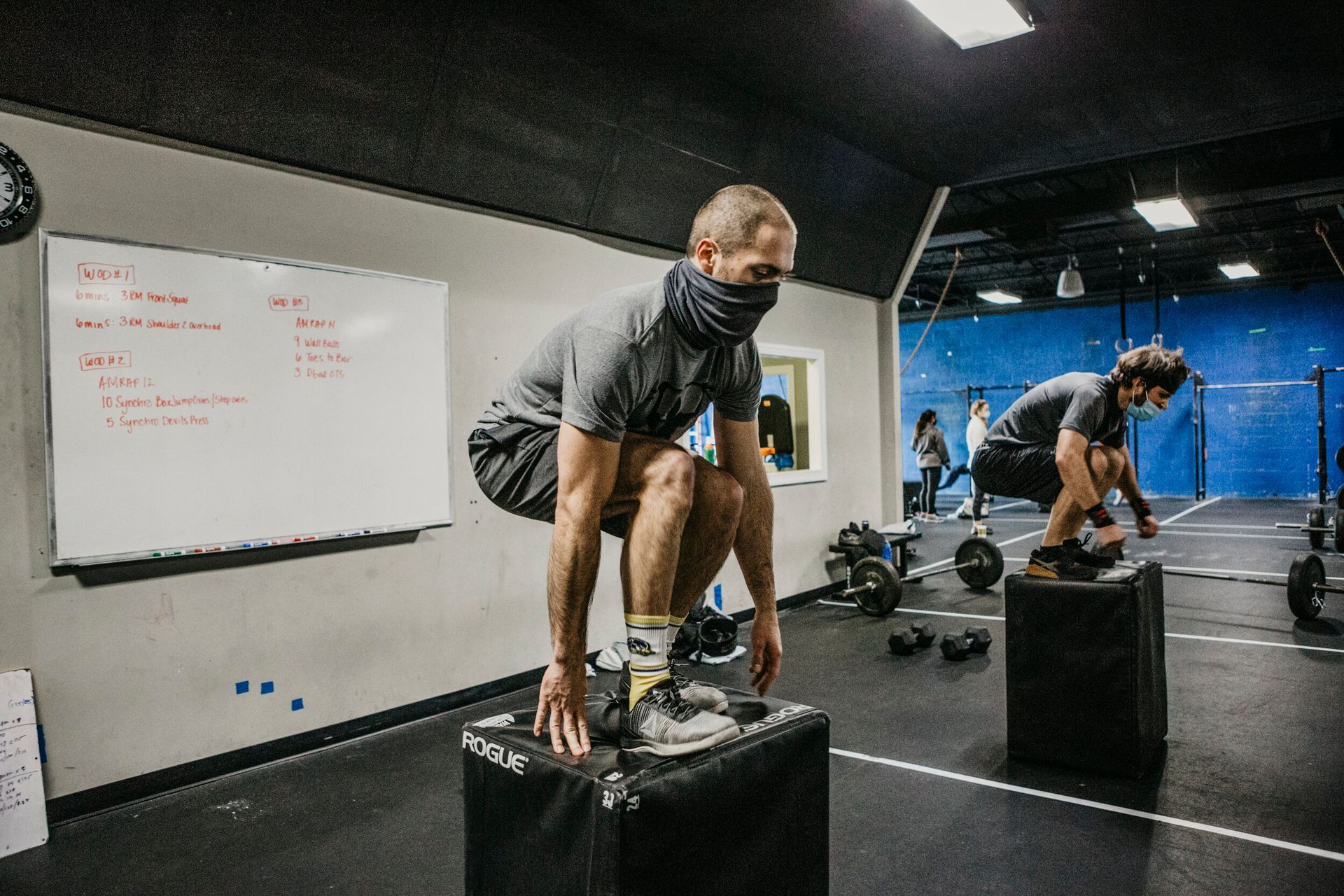 A man wearing a mask is squatting on a box in a gym.