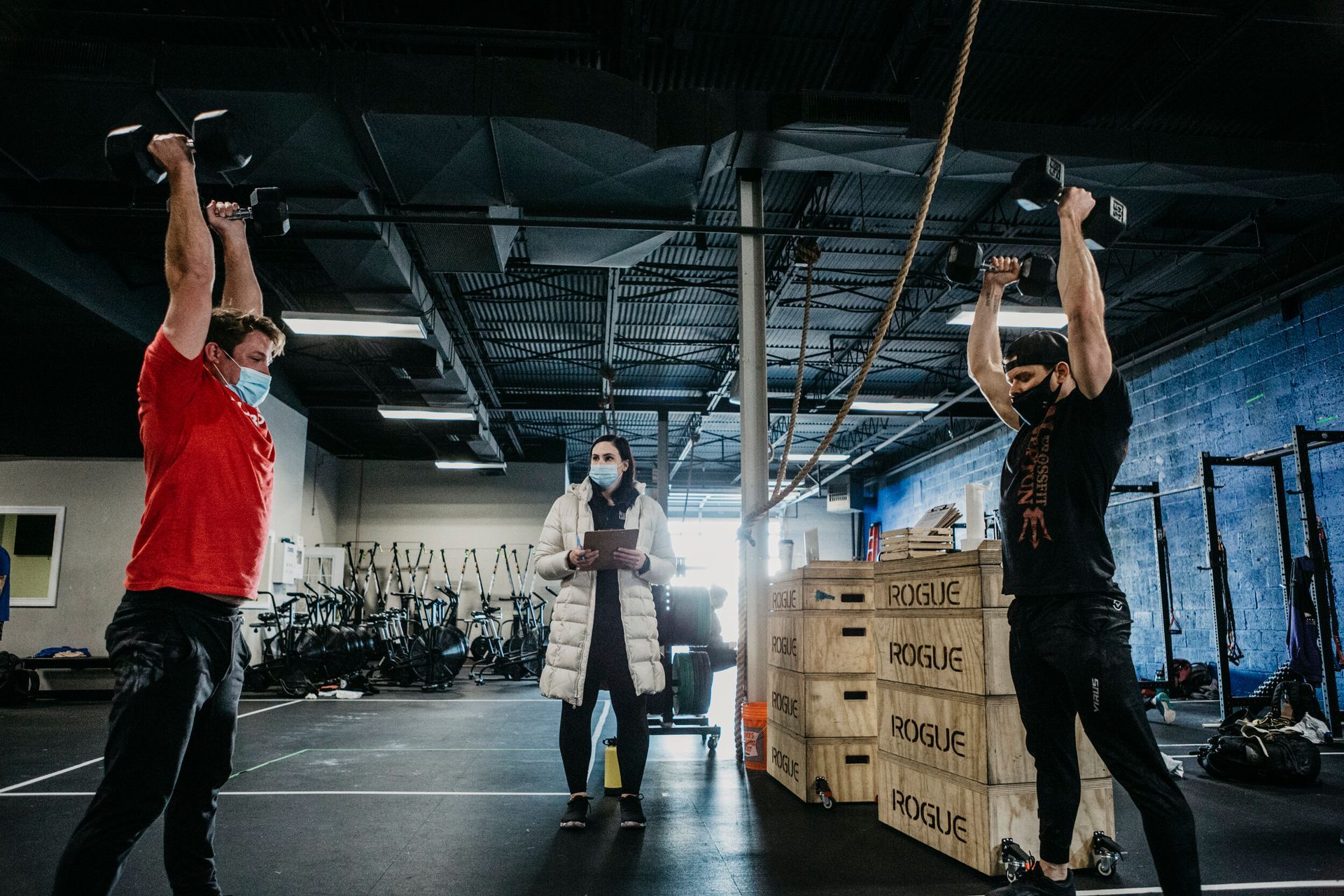 A man is lifting a dumbbell over his head in a gym while a woman watches.