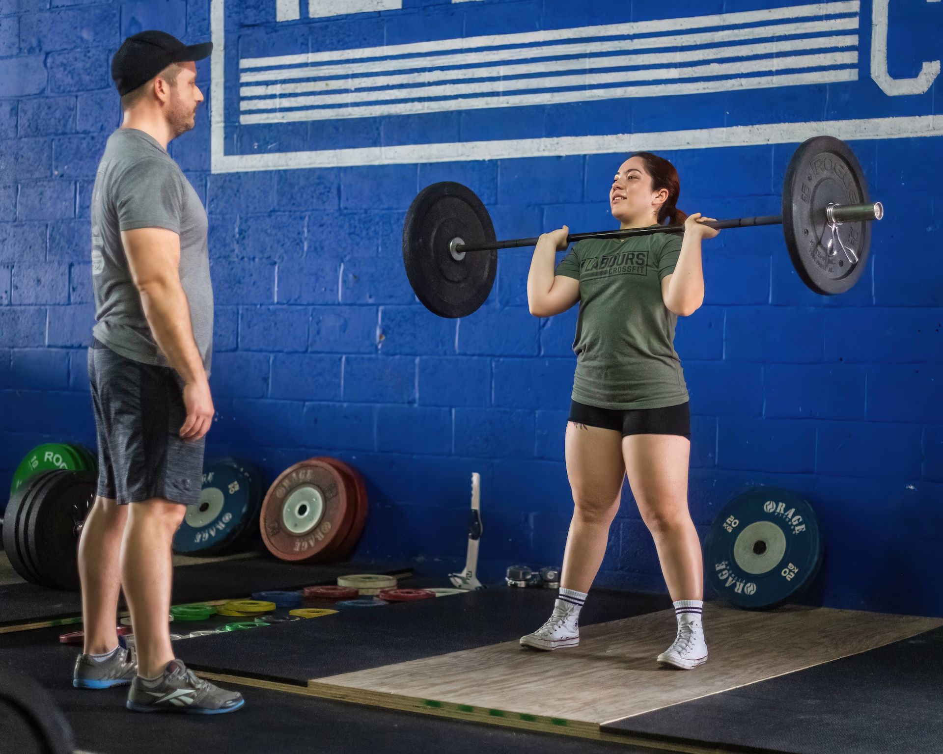 A man is coaching a member lifting a barbell in a gym.