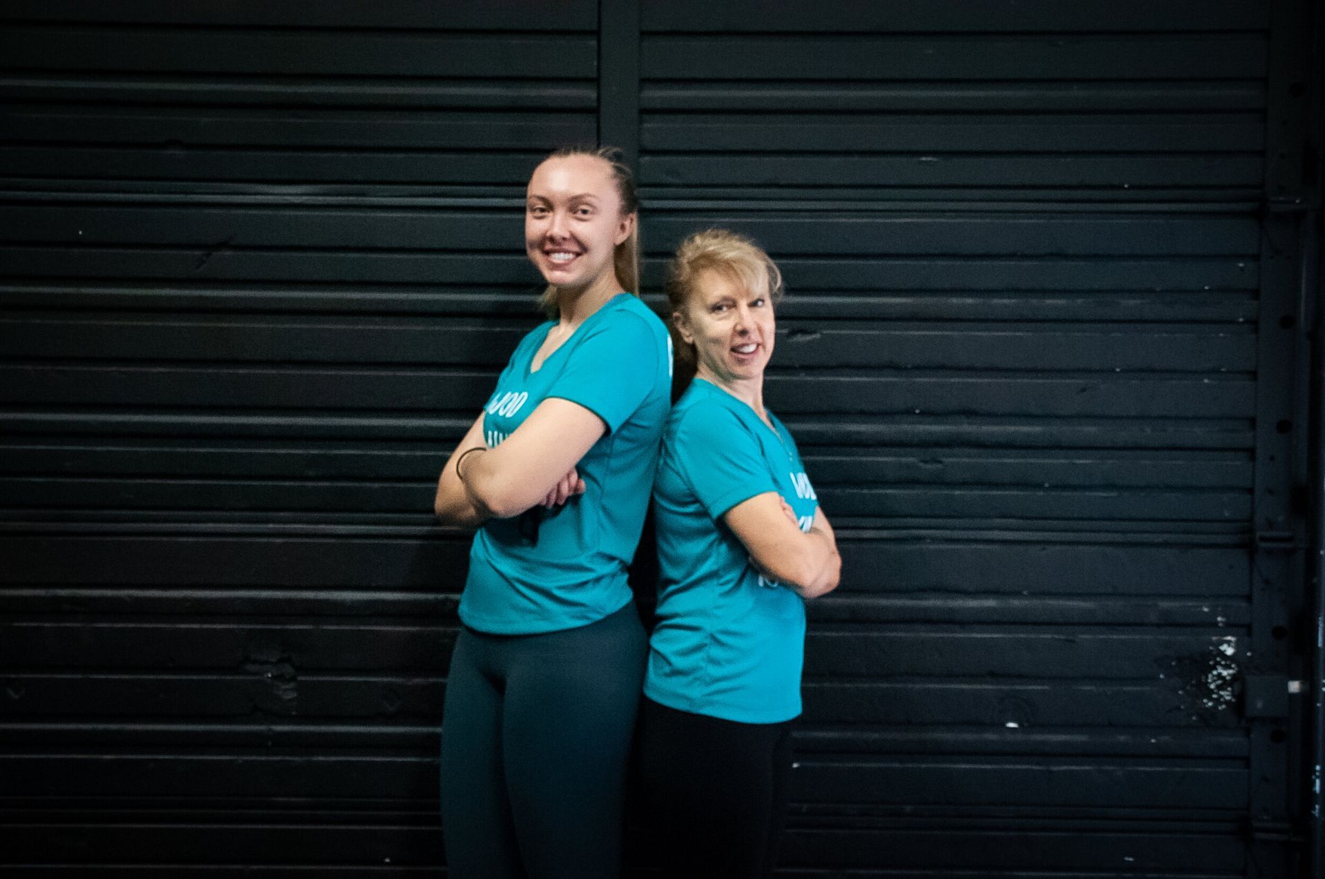 Two women are standing back to back in front of a black wall.